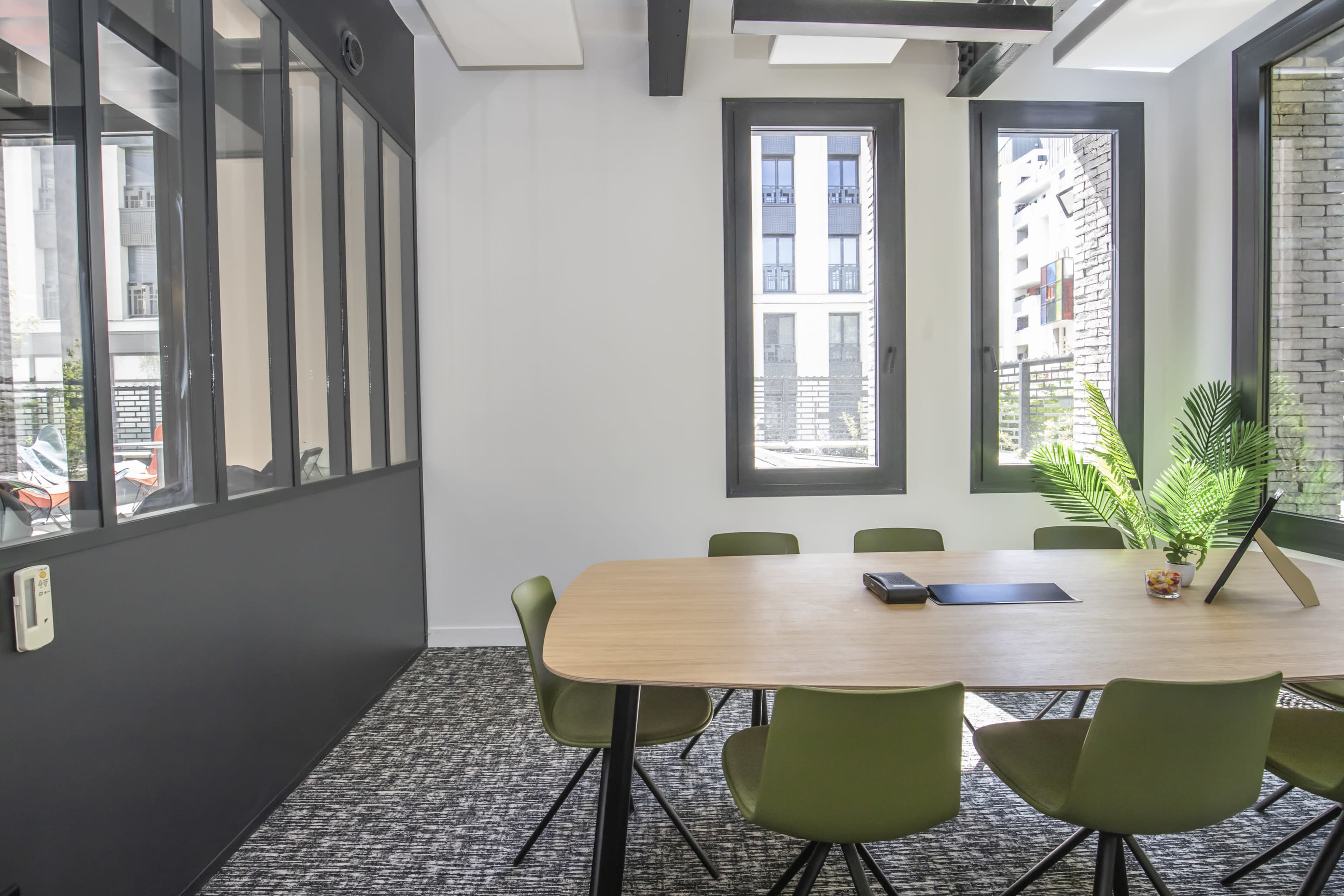A modern conference room features a wooden table surrounded by green chairs, with large windows allowing natural light to enter.