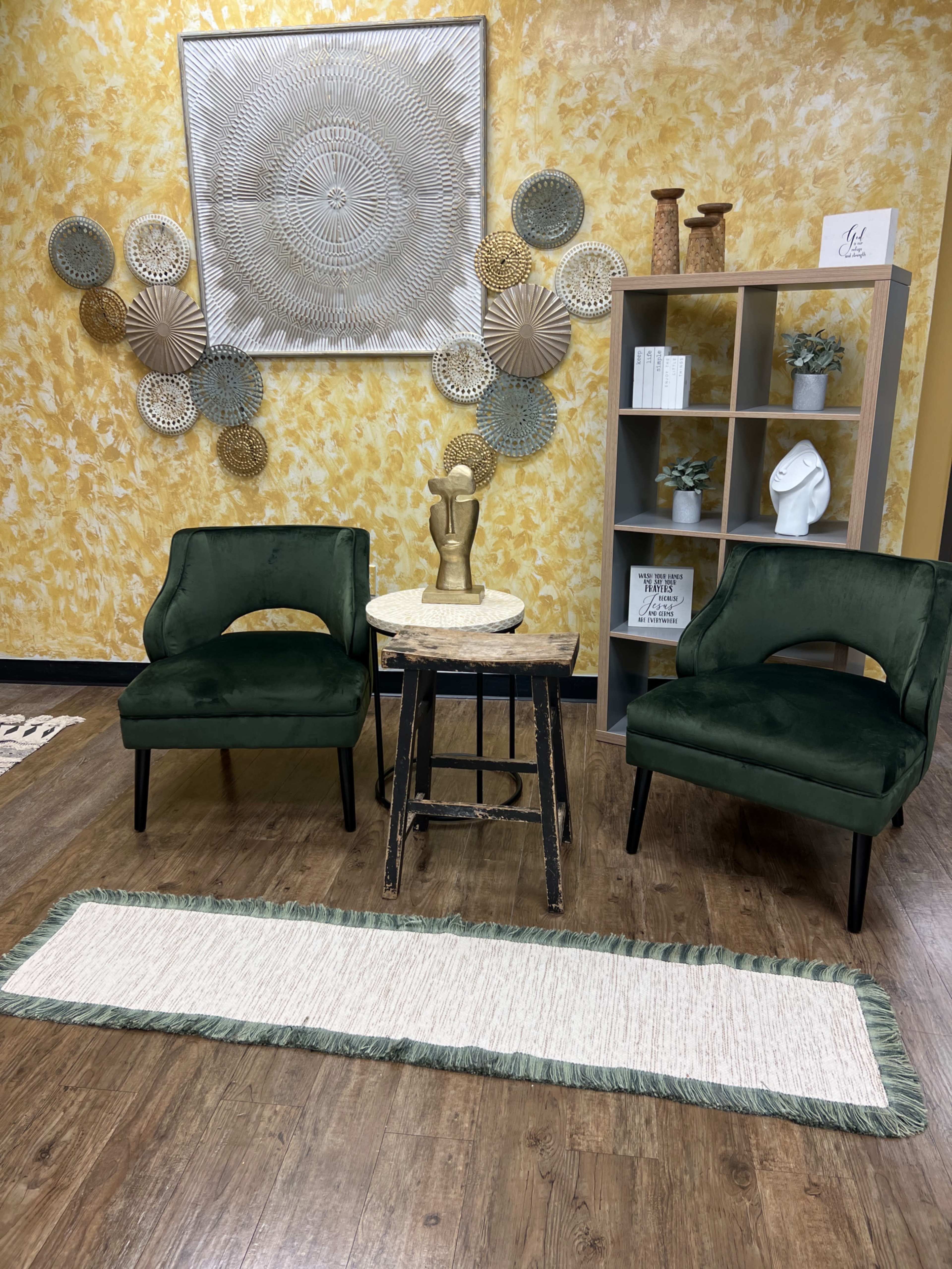 The image showcases a small seating area with two green chairs, a round table with a decorative sculptural piece, a patterned wall decoration, and a light-colored runner on the wooden floor.