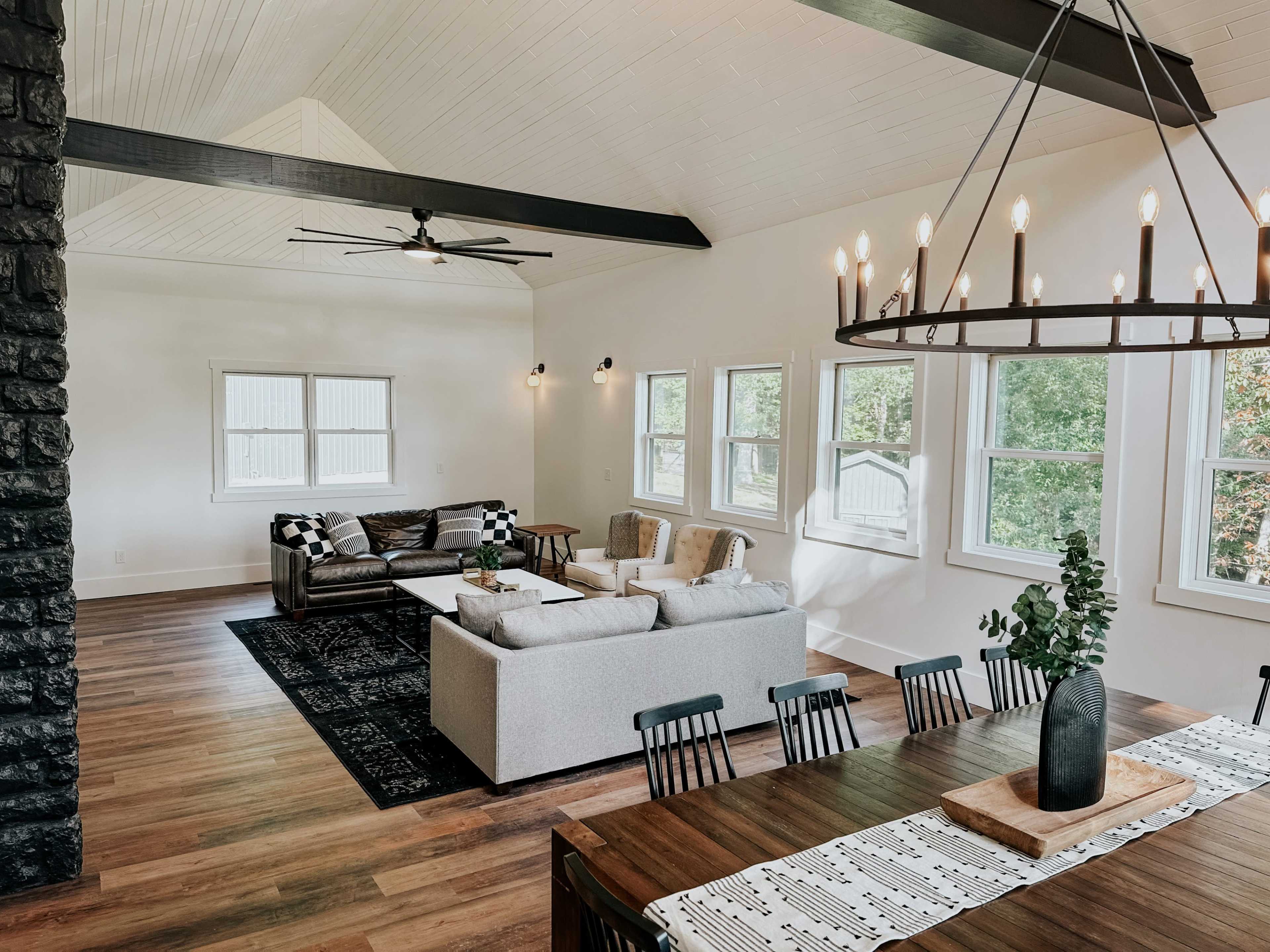 A spacious living area features a mix of modern furniture, large windows, and a dining table set in a well-lit room with wooden flooring.