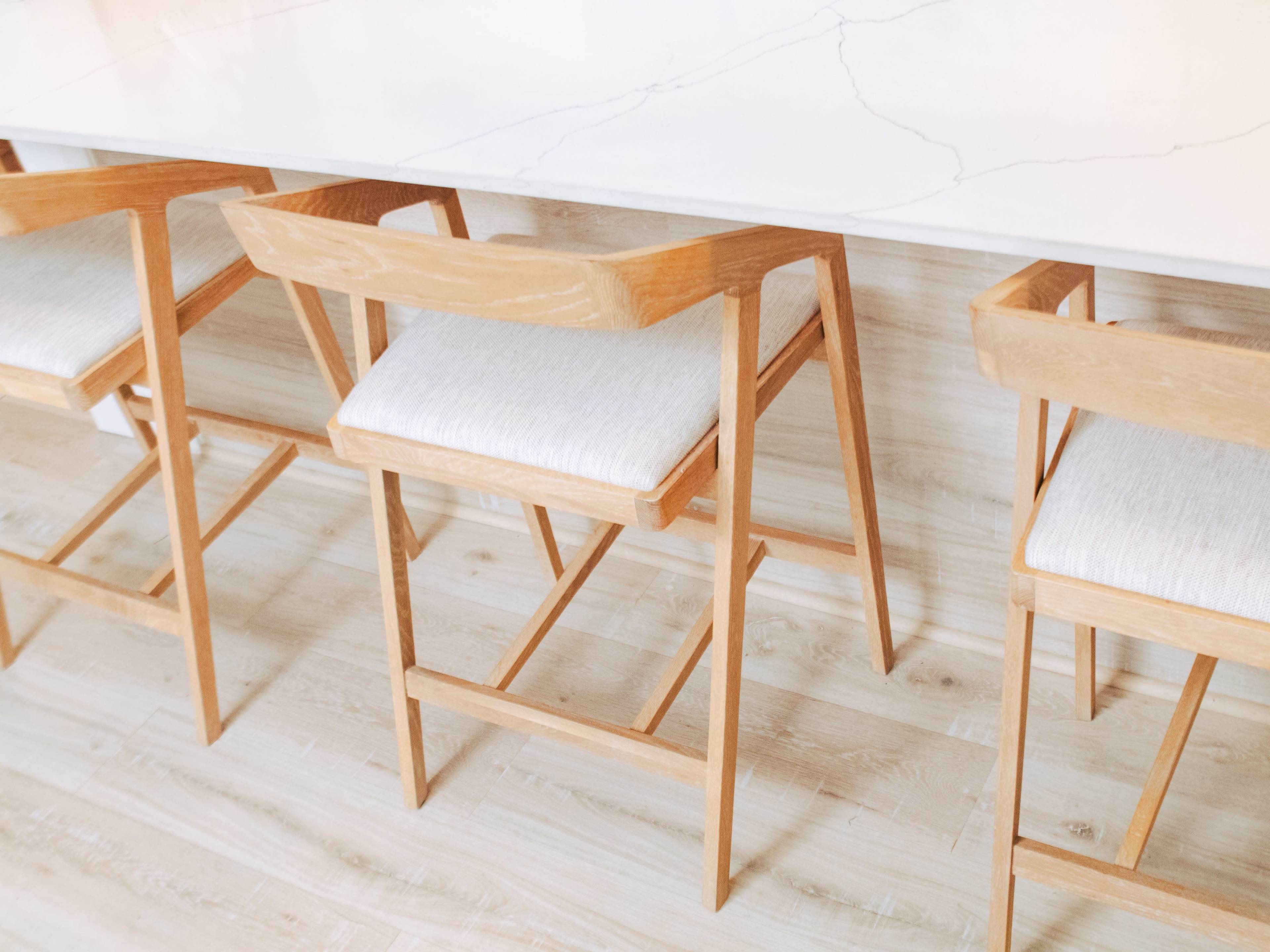 Three wooden bar stools with light-colored fabric cushions are positioned around a marble-topped table on a wooden floor.