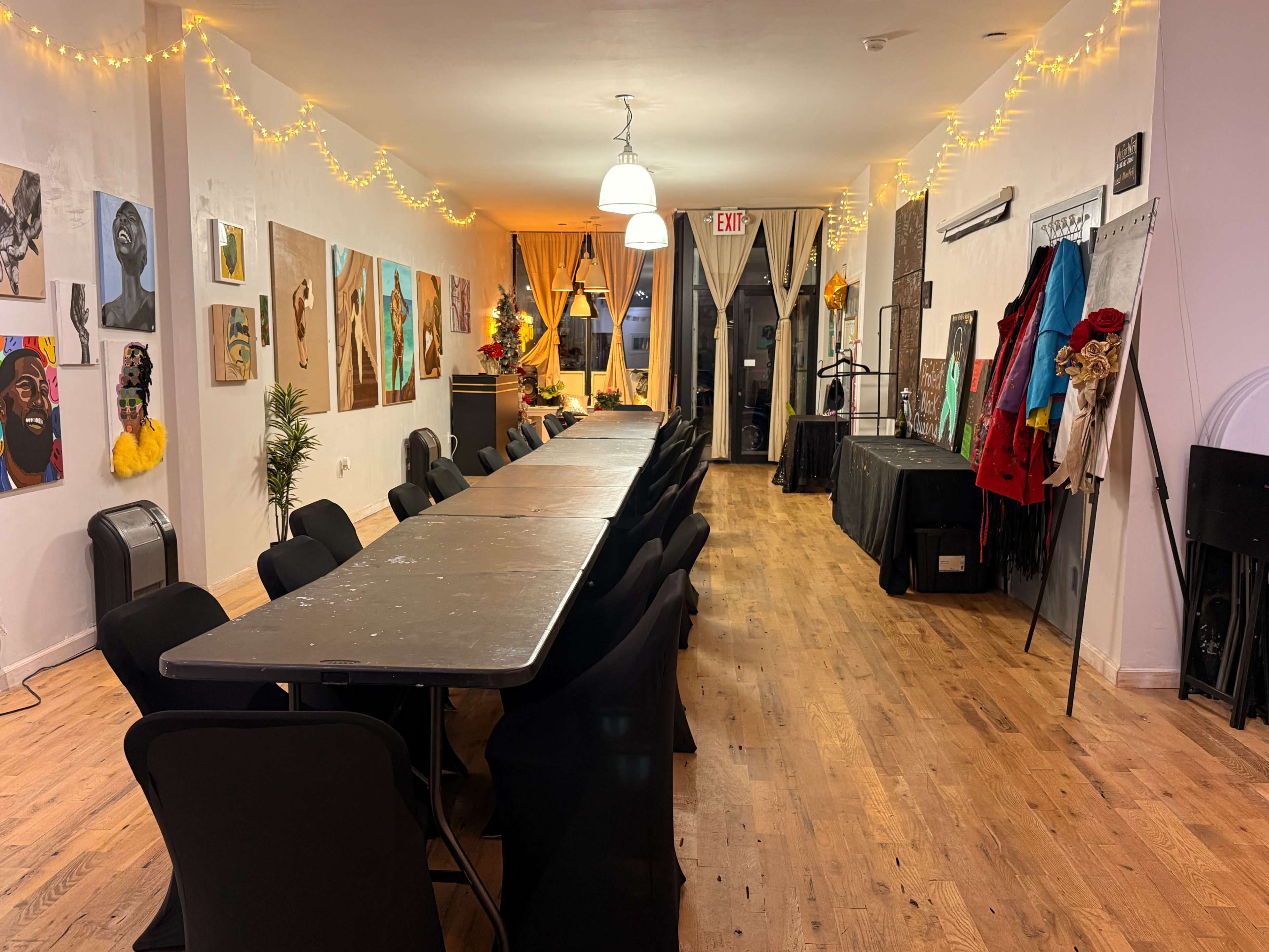 The image shows a well-lit indoor space with a long table surrounded by chairs, decorated with various art pieces on the walls and string lights overhead.