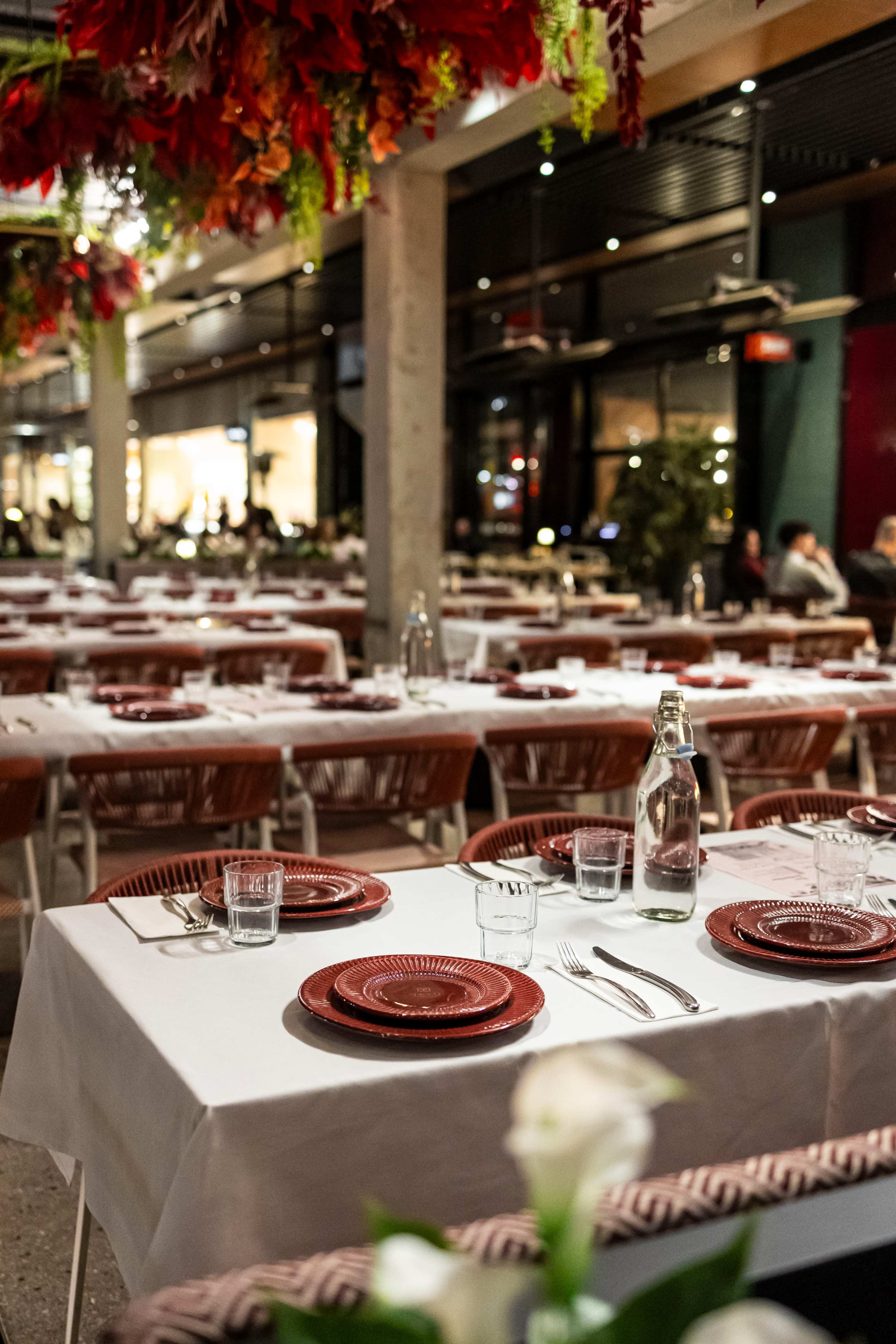 The image shows a dining area with multiple tables set with red plates and glassware, surrounded by decorative greenery hanging from the ceiling.