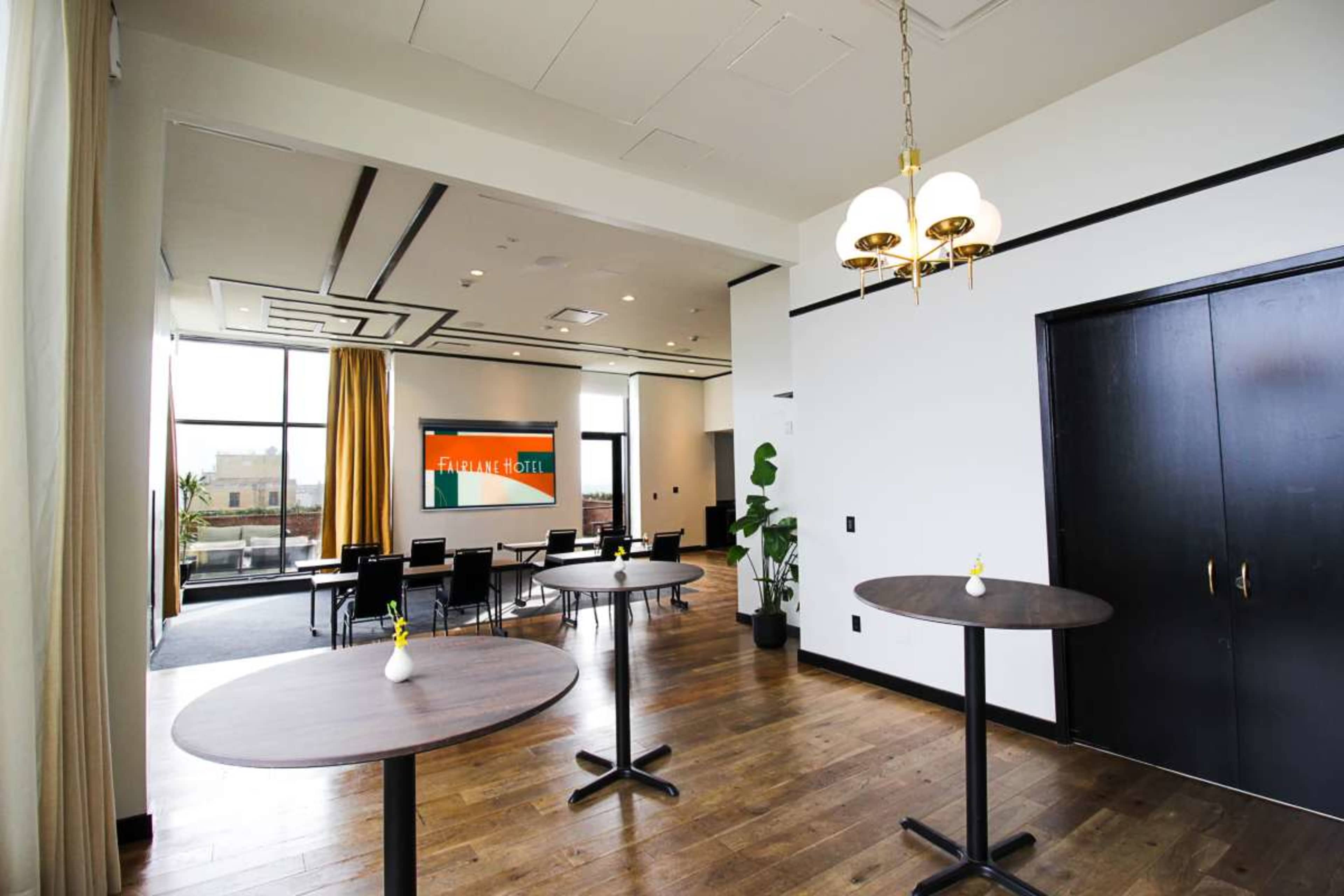 The image shows a modern event space with tall windows, two round tables with decorative vases, and a large screen displaying an image on the wall.