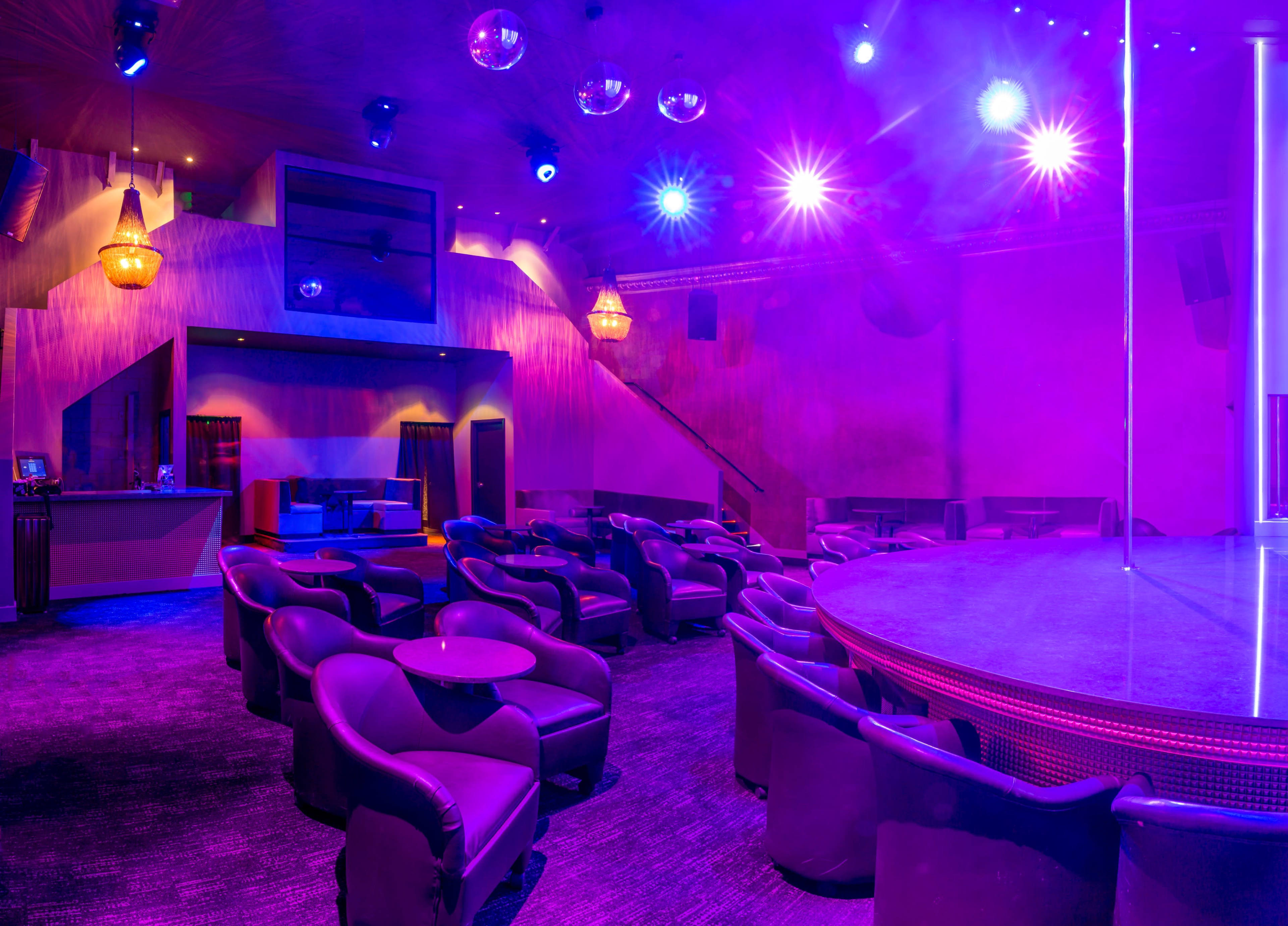 The image shows a dimly lit club interior featuring a circular stage, plush seating, and colorful lights.
