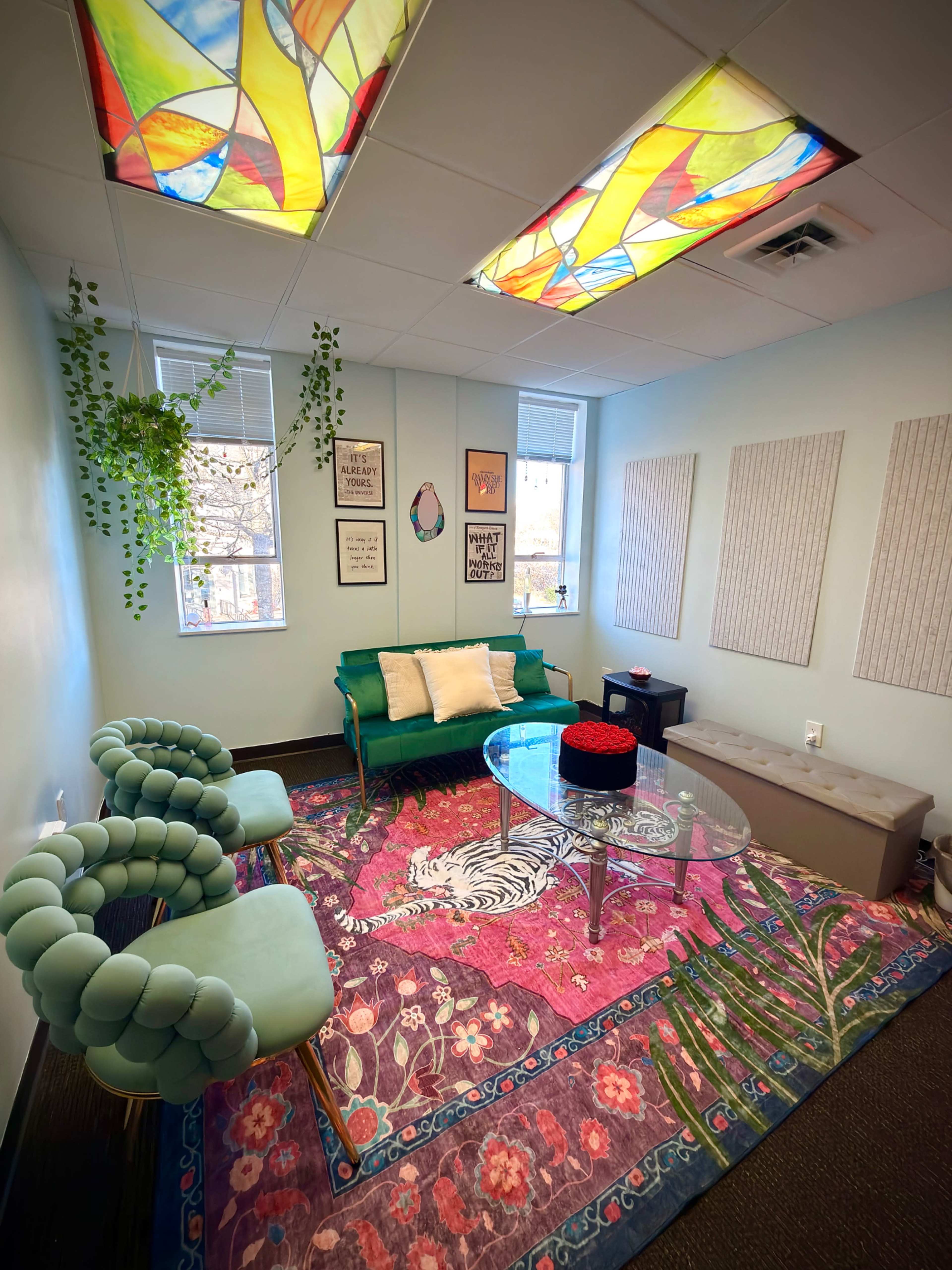 The image shows a softly lit room featuring two green chairs, a glass coffee table, colorful wall decor, and a large, vibrant rug with floral patterns.