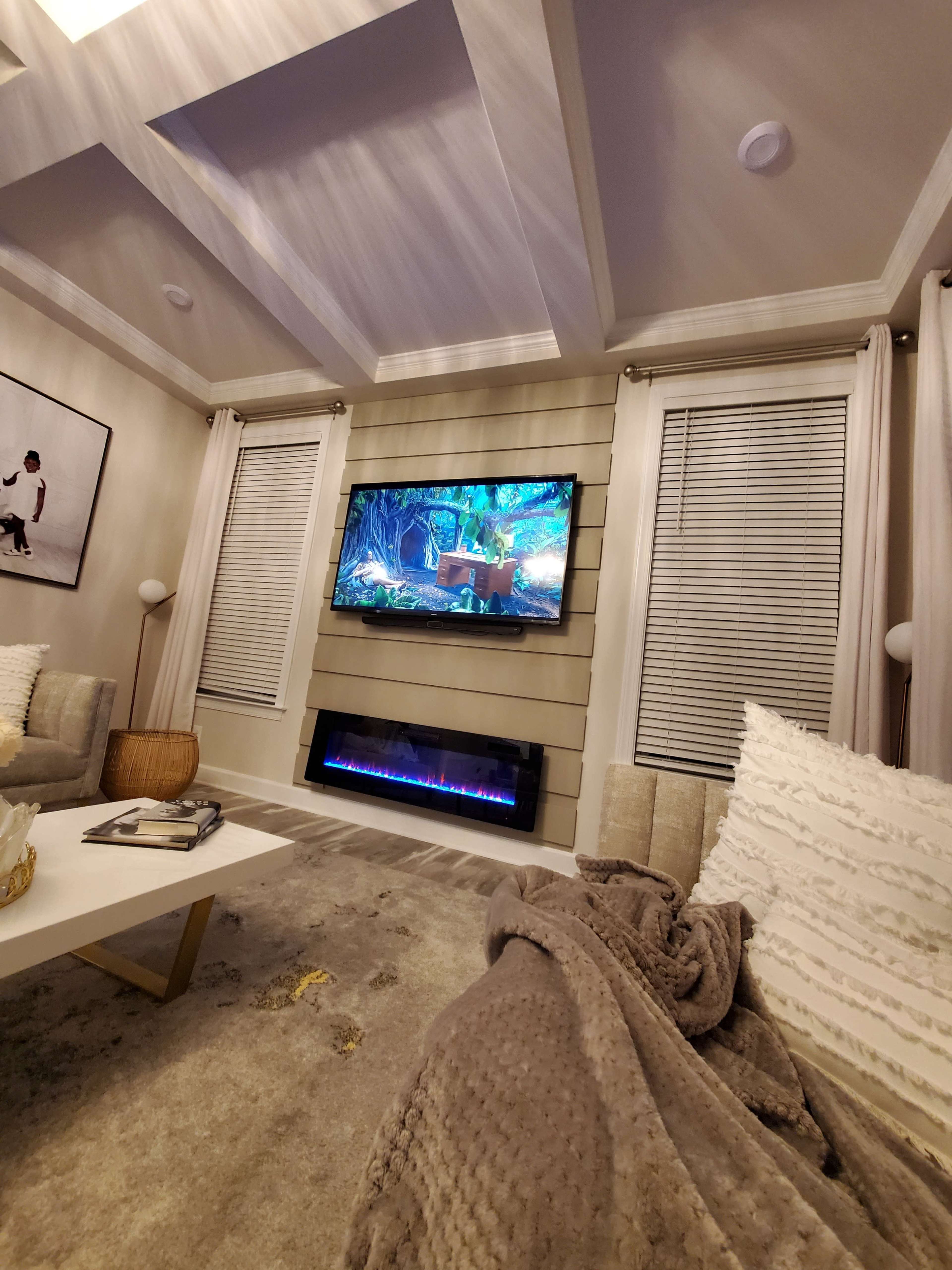 The image shows a modern living room with a wall-mounted television, a cozy electric fireplace, and two plush sofas, all illuminated by ceiling lights.