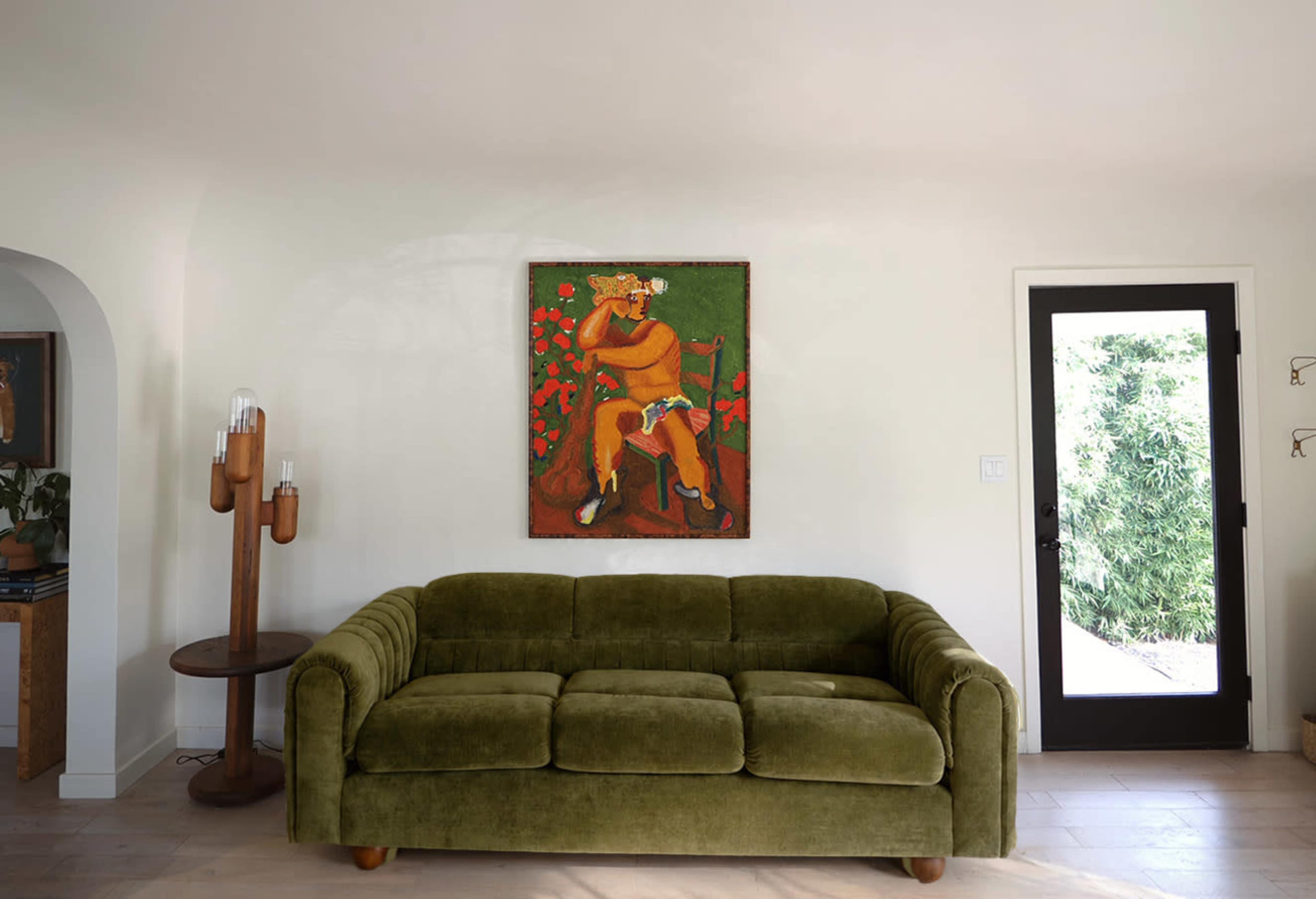 The image shows a green, plush couch facing a wall with a colorful painting of a seated figure against a backdrop of red flowers, next to a door opening to an outdoor area.