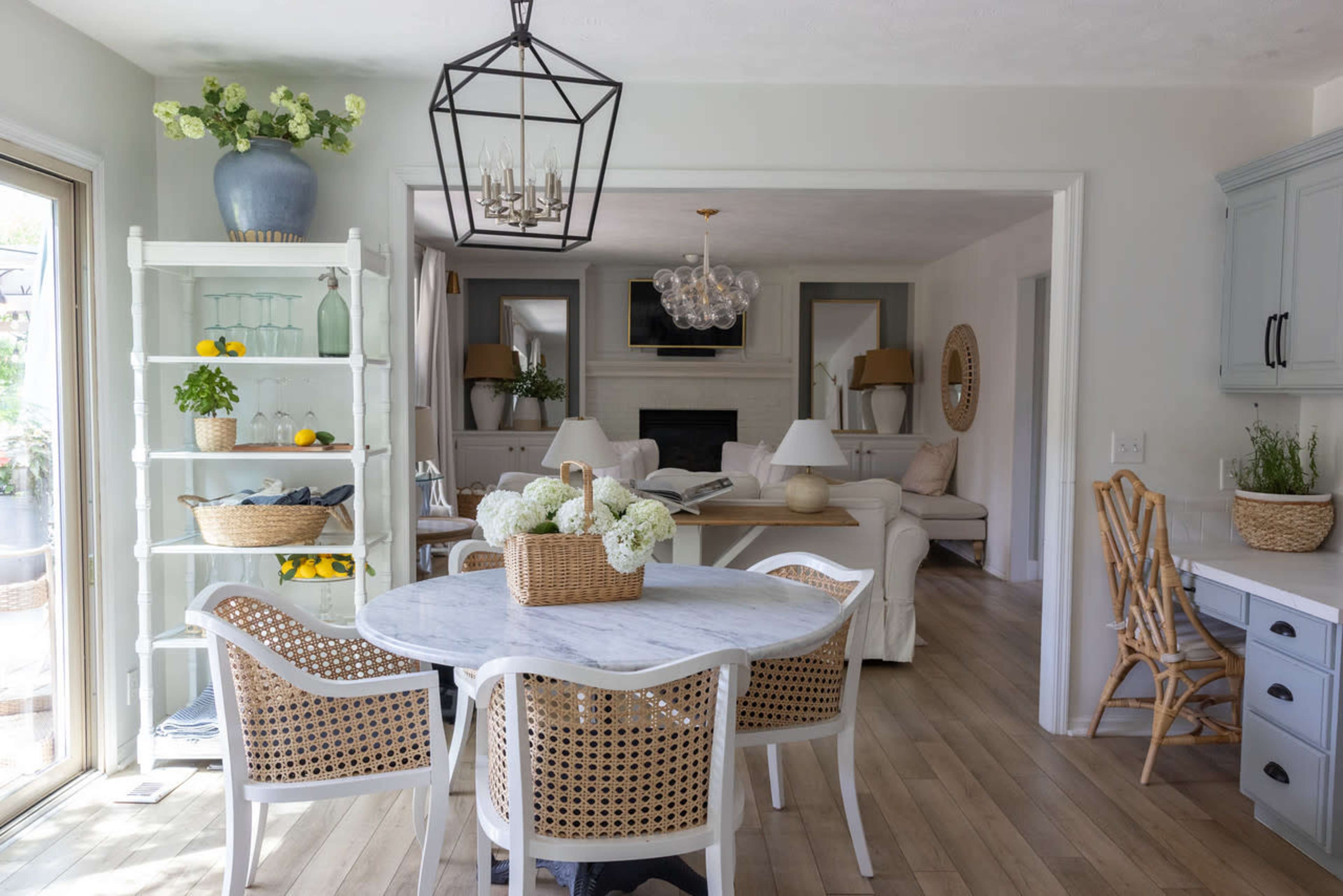 A light-filled dining area features a round table surrounded by white and rattan chairs, with a bookshelf and a living room in the background.
