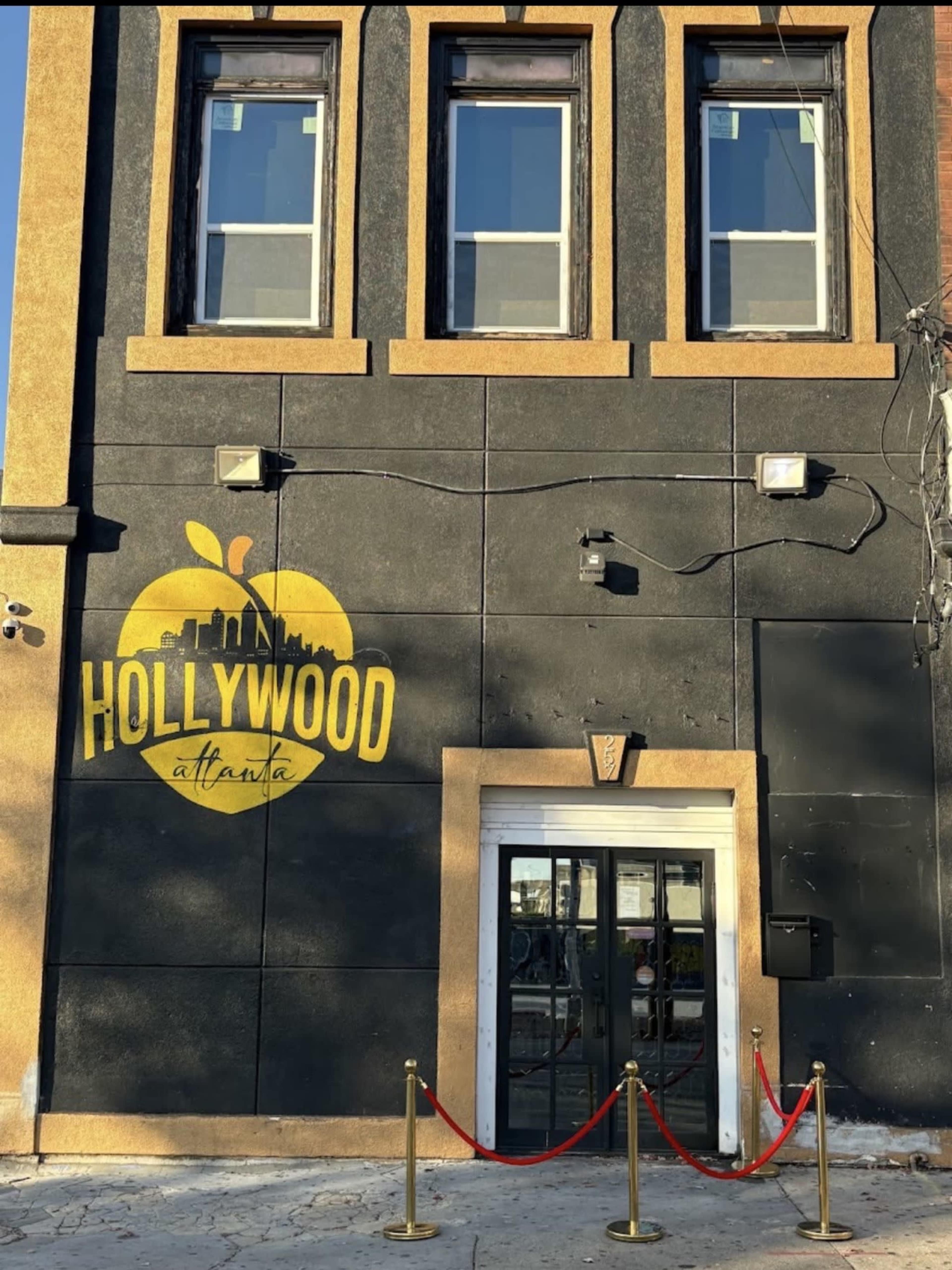 The image shows the exterior of a building with a large yellow logo that reads "HOLLYWOOD" and "atlanta," featuring a red carpet and stanchions in front of the entrance.