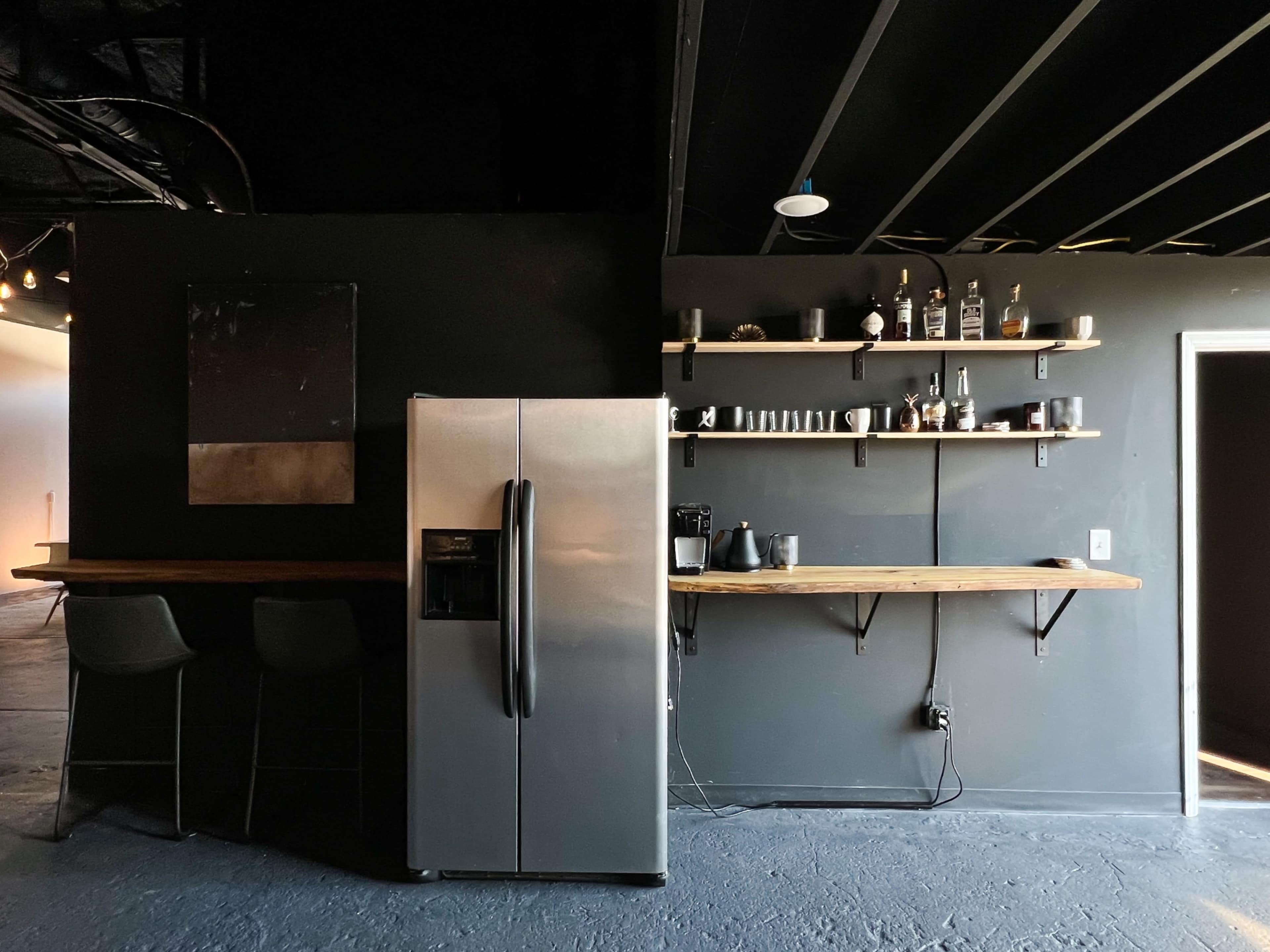 A modern kitchen area featuring a stainless steel refrigerator, a wooden bar counter with stools, and shelves displaying various glassware and appliances against a dark wall.
