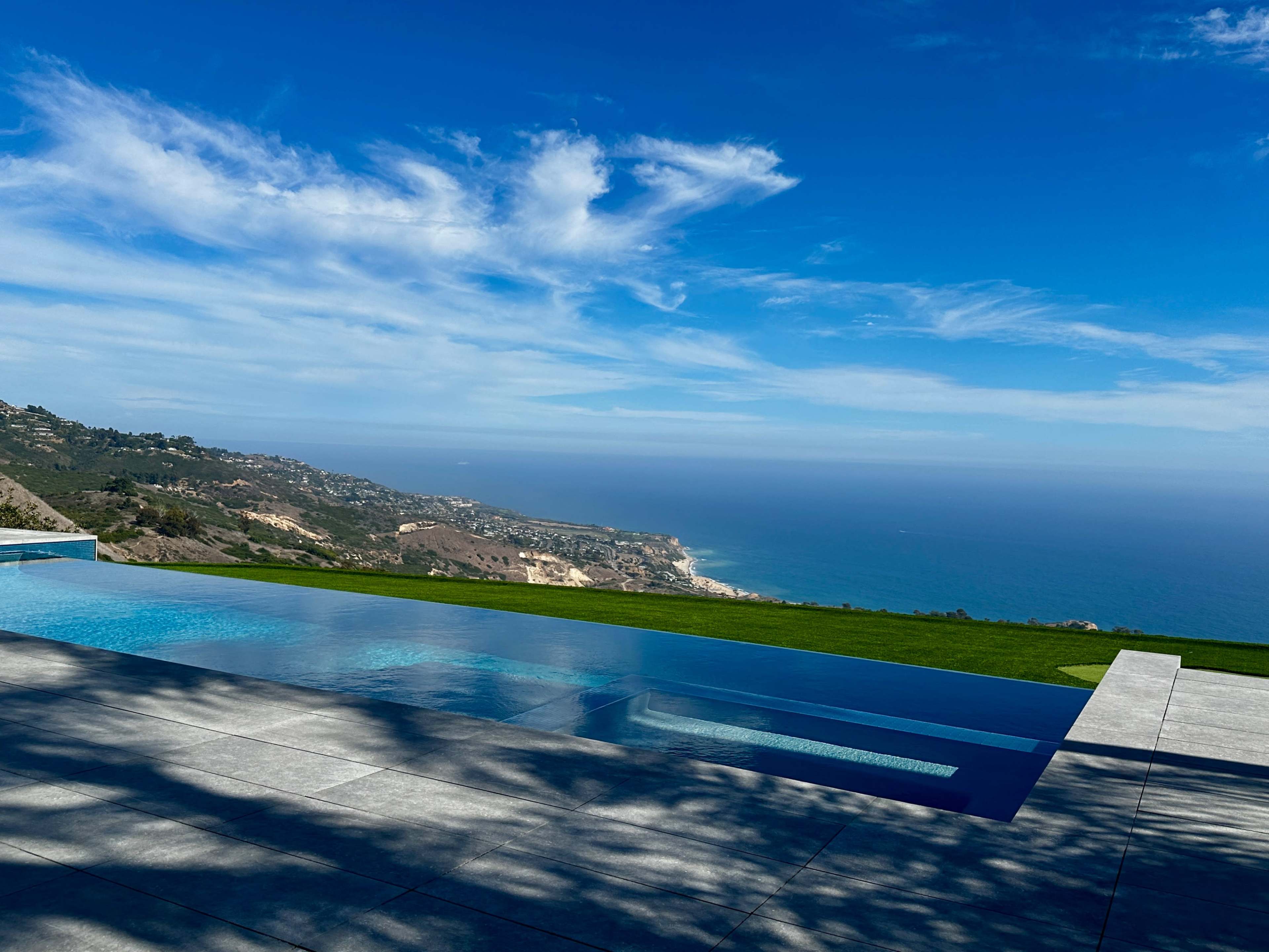 A modern infinity pool overlooks a scenic coastal landscape under a clear blue sky.