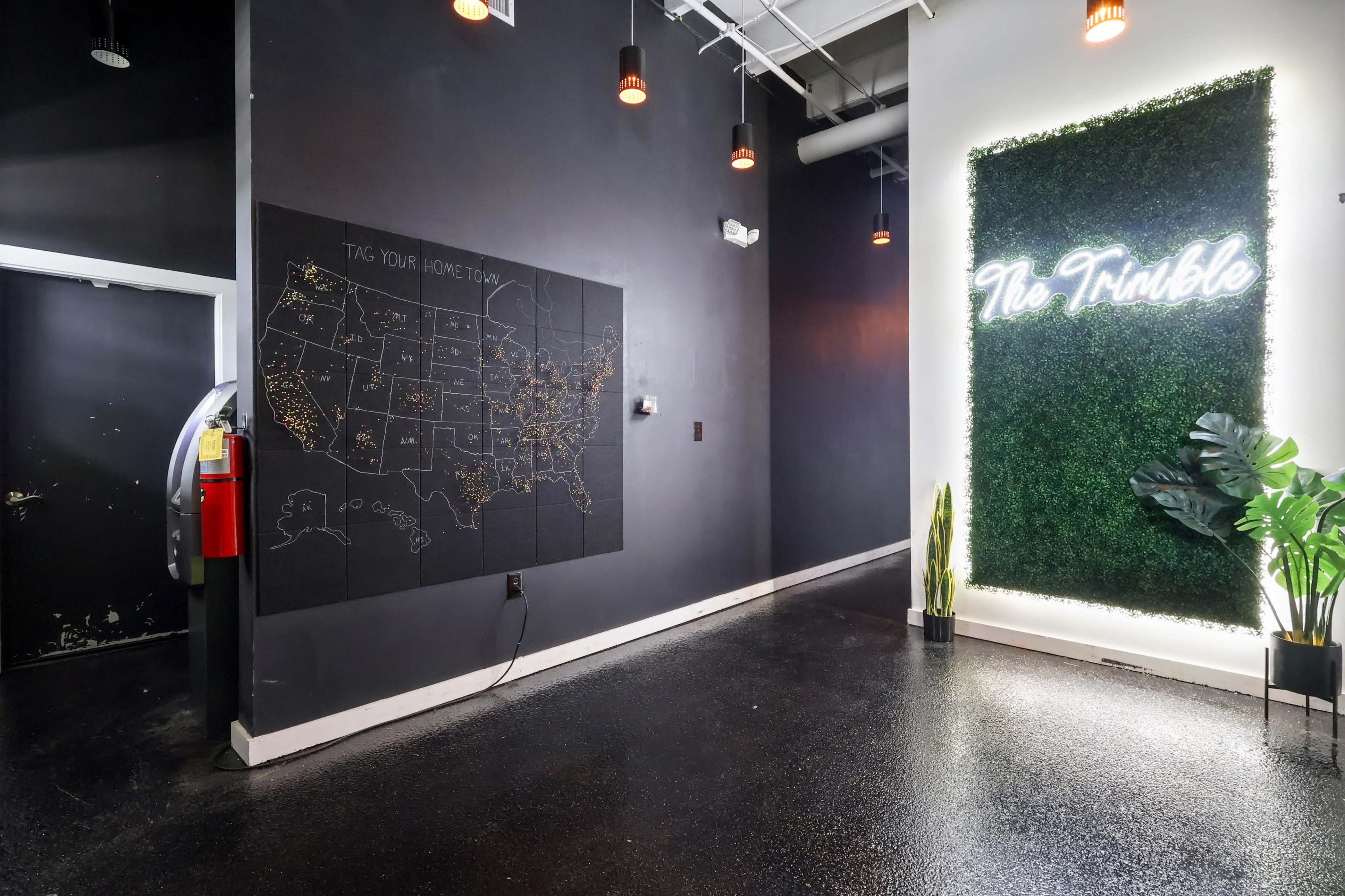 The image shows a modern interior space featuring a wall with a dark chalkboard map of the United States and a lighted sign that reads "The Trimble" next to a green wall with plants.