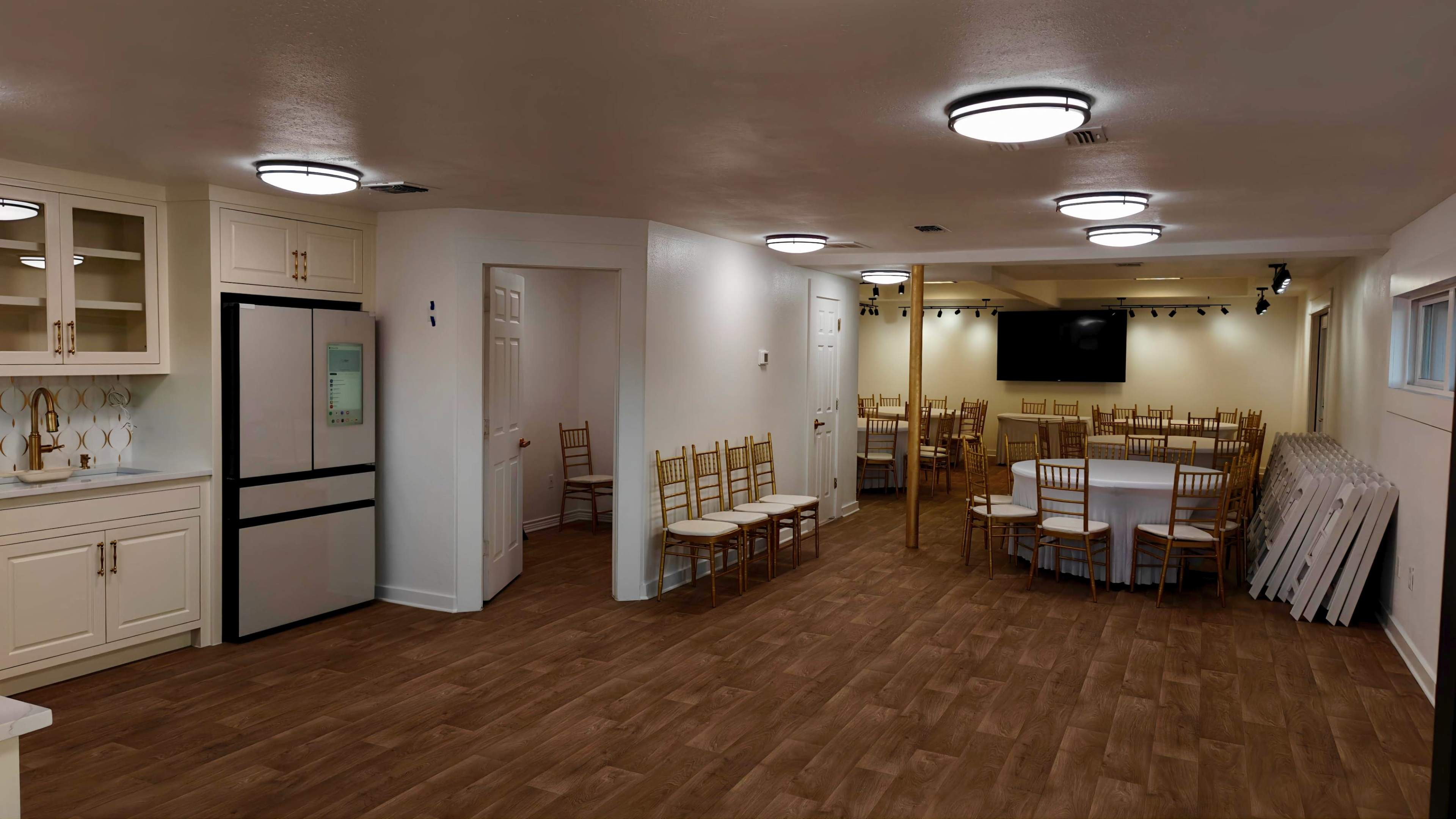 The image shows a spacious room with tables and chairs arranged for seating, a kitchen area at one end, and a large screen at the far wall.