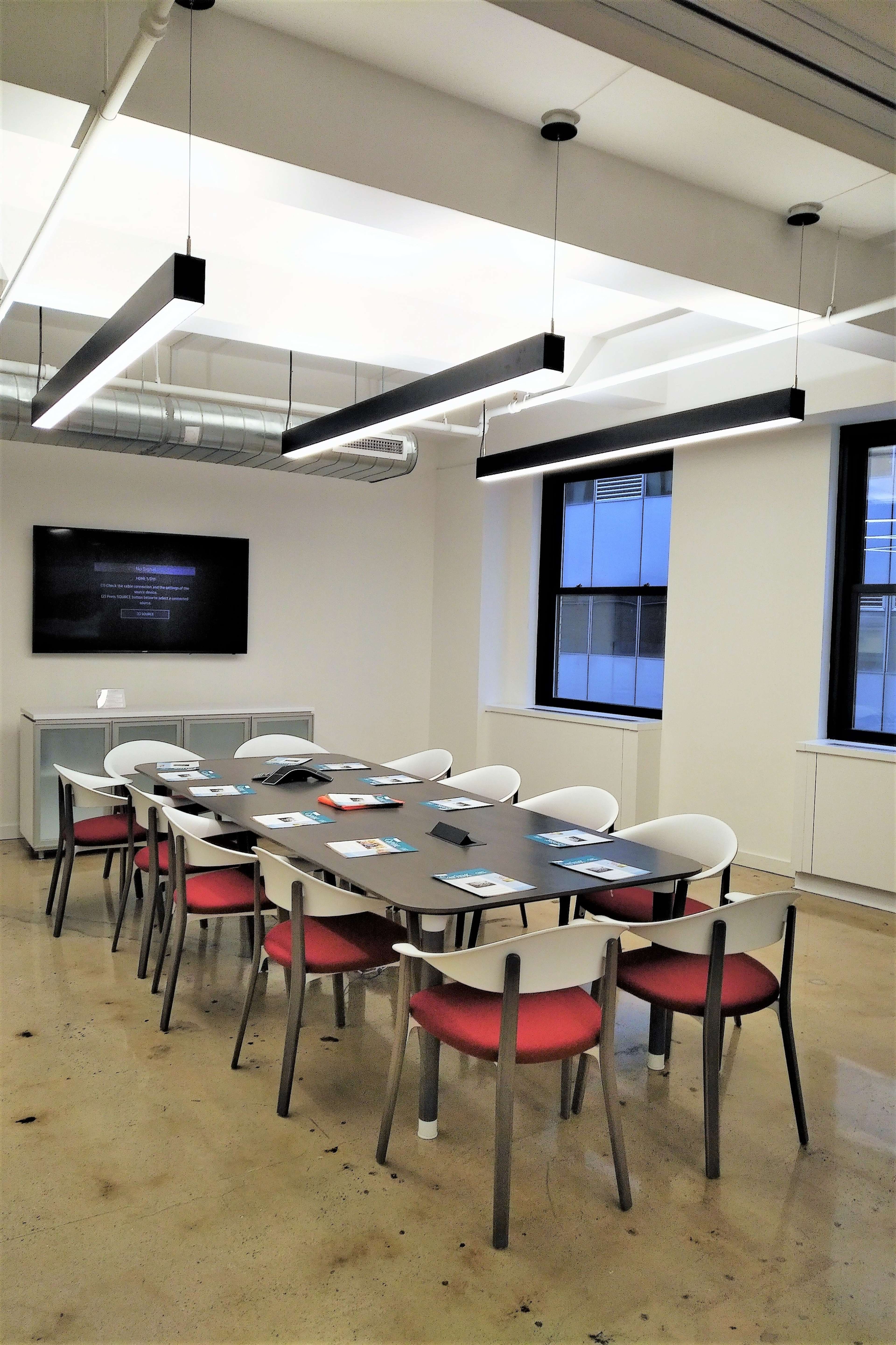 A modern conference room contains a long table surrounded by chairs, with a large screen on one wall and overhead lighting.