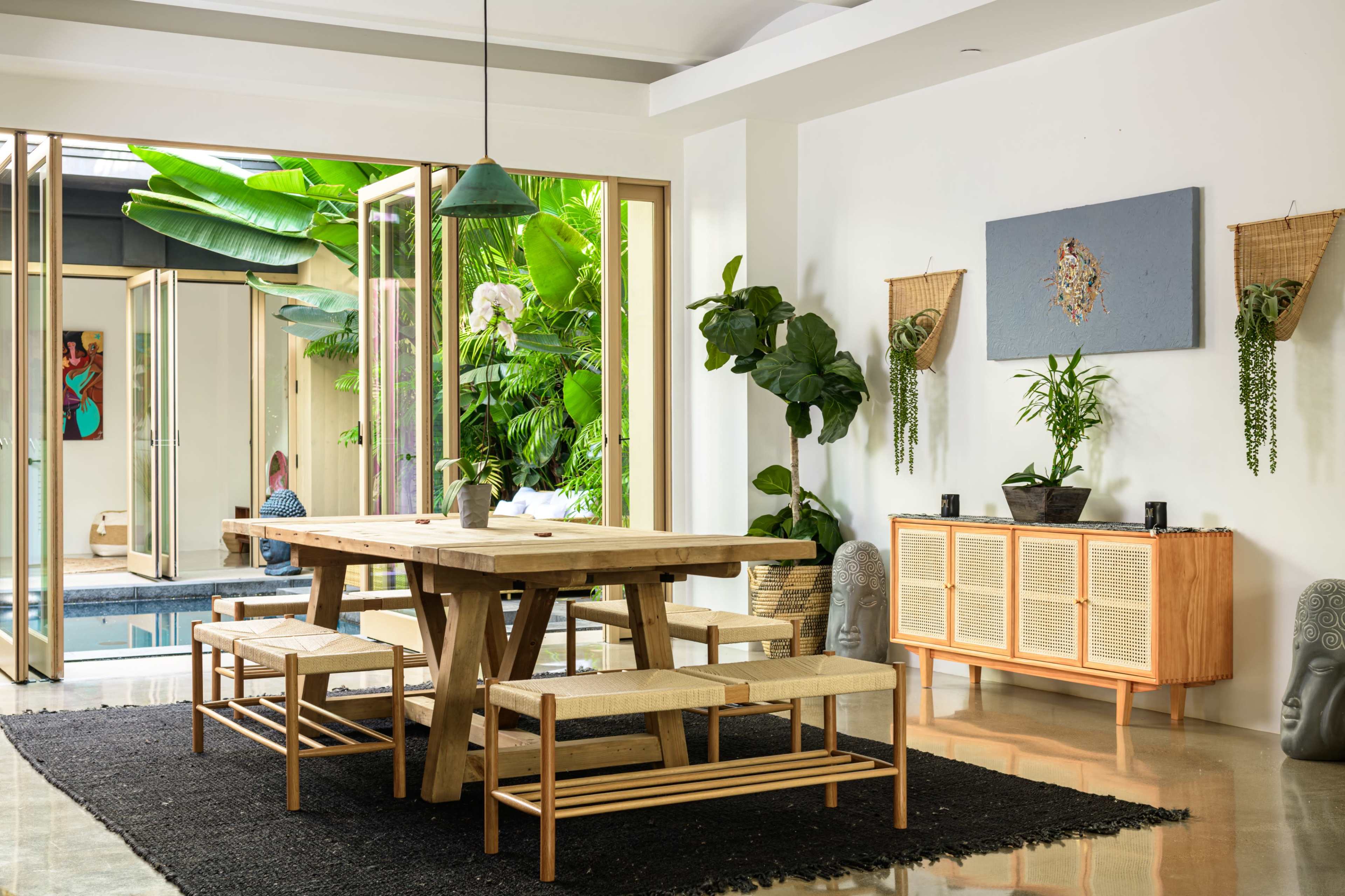 A dining area features a large wooden table with benches, surrounded by plants and opening to a poolside view through glass doors.