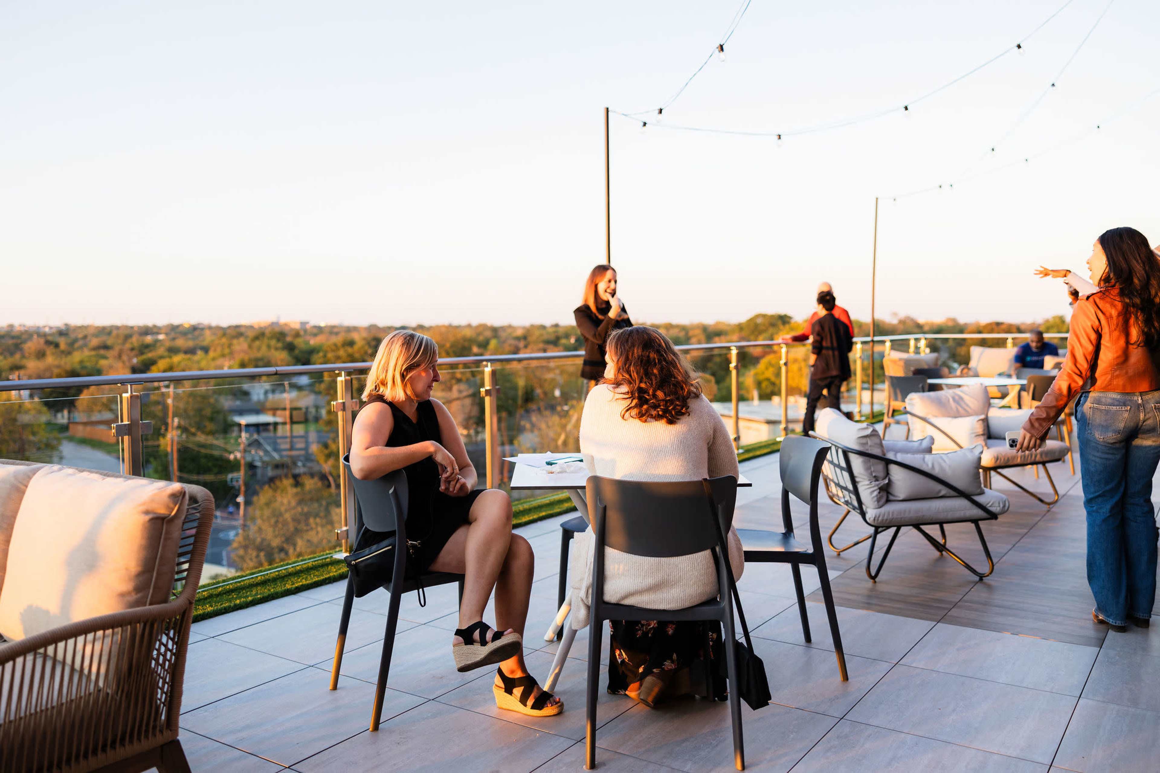 A rooftop gathering features several people chatting and enjoying the view at sunset, with modern seating arranged around them.