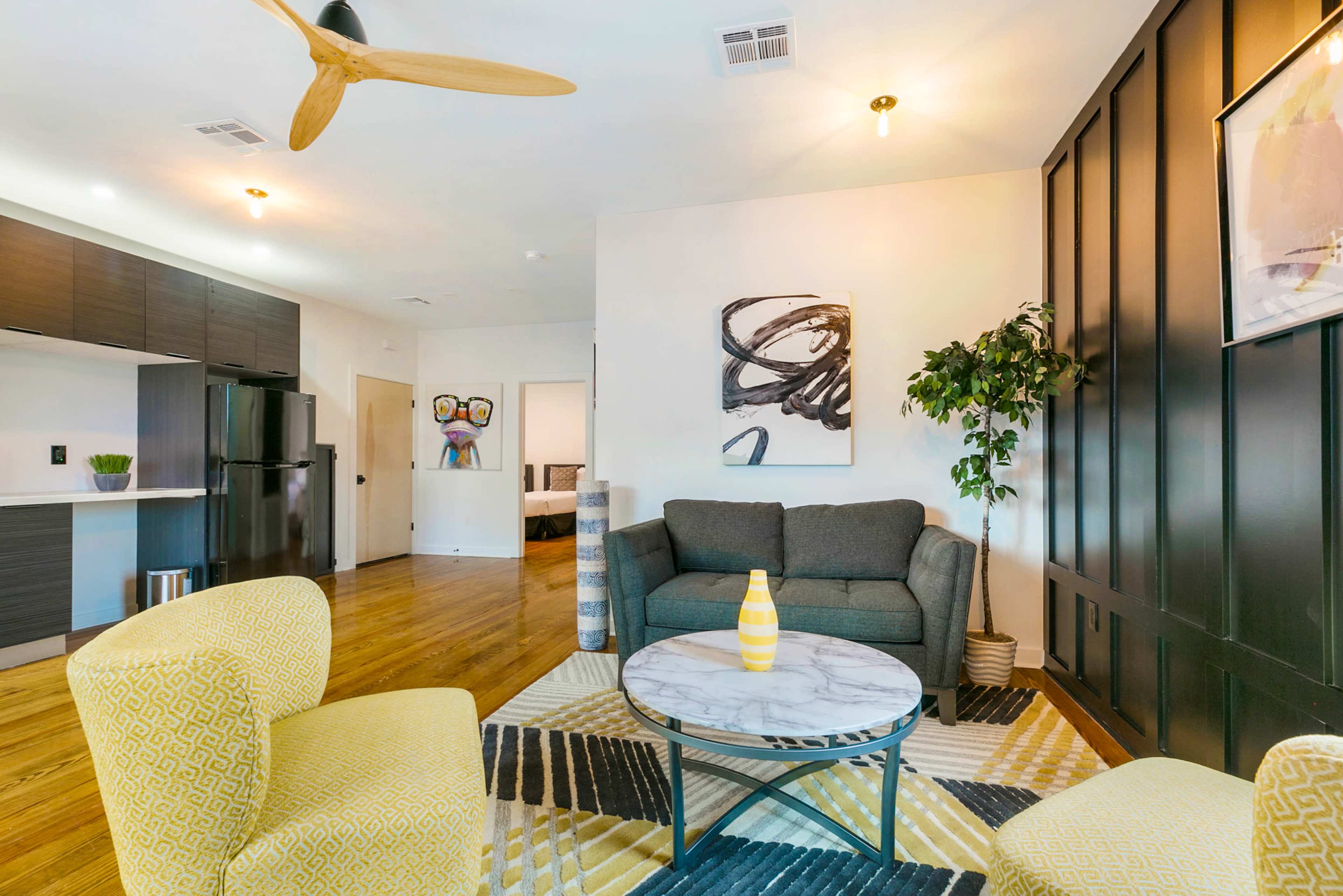 A modern living room features a gray sofa, two yellow patterned chairs, a marble coffee table, and a wall art piece, with a ceiling fan and wood flooring.