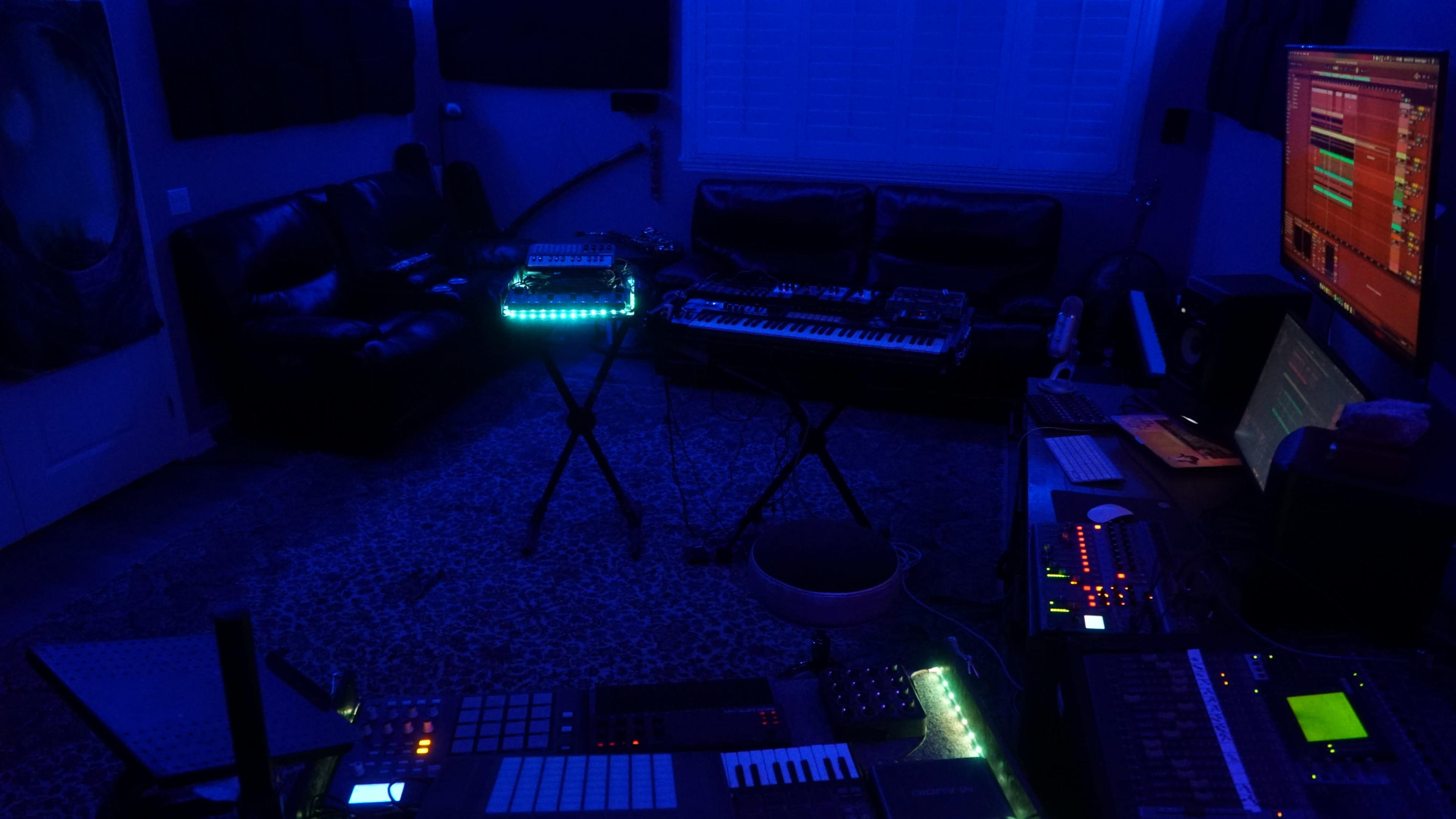 A dimly lit music studio features electronic instruments arranged on stands with soft blue lighting and a couch in the background.