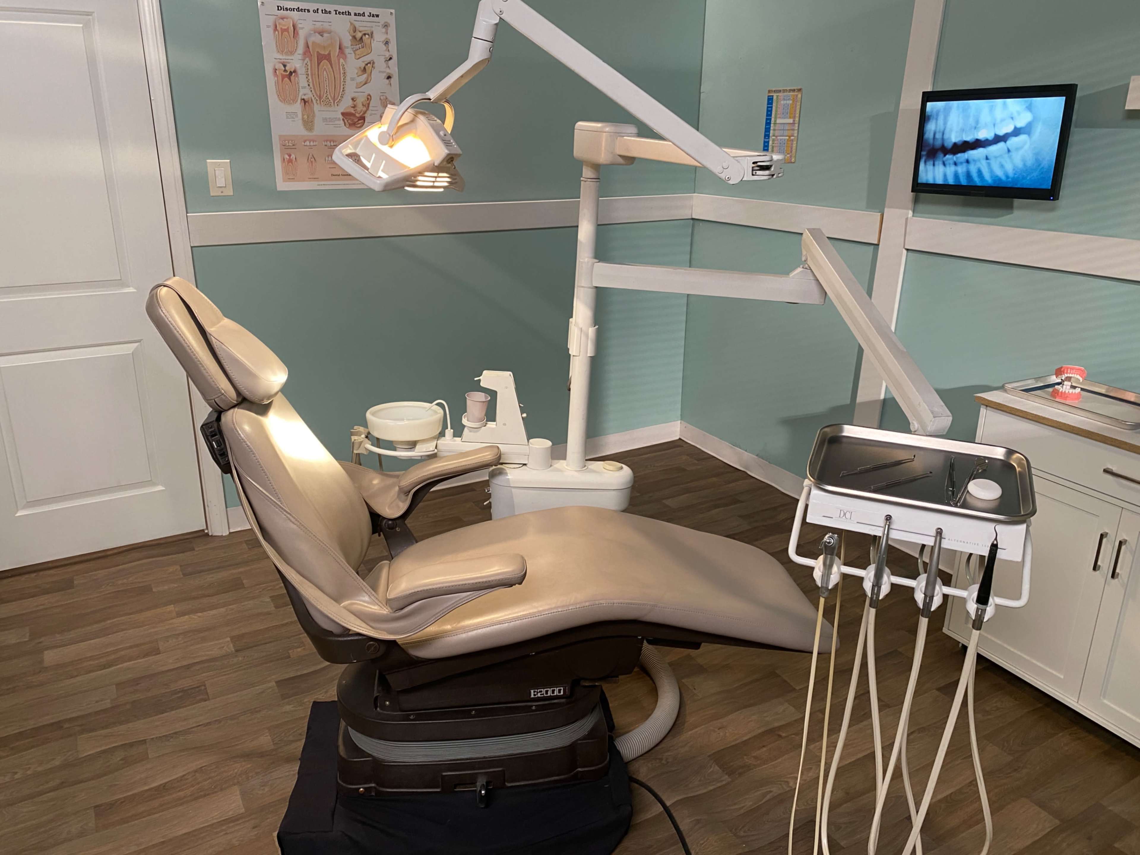 The image shows a dental examination room featuring a dental chair, overhead light, and an X-ray viewing monitor on the wall.