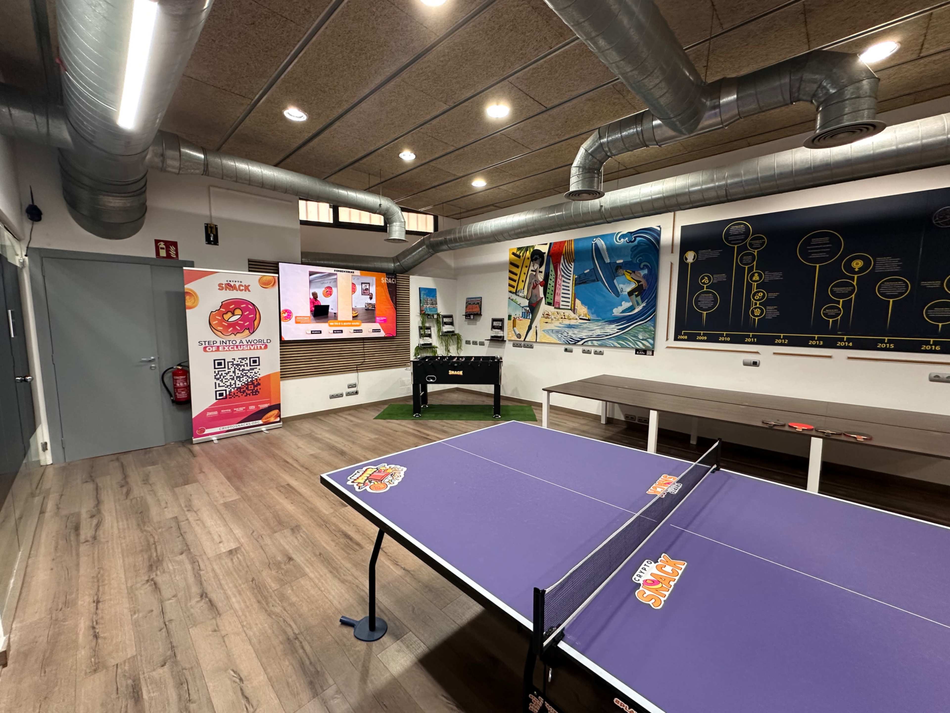 The image shows a modern recreational room with a ping pong table, a promotional banner, and walls decorated with colorful artwork.