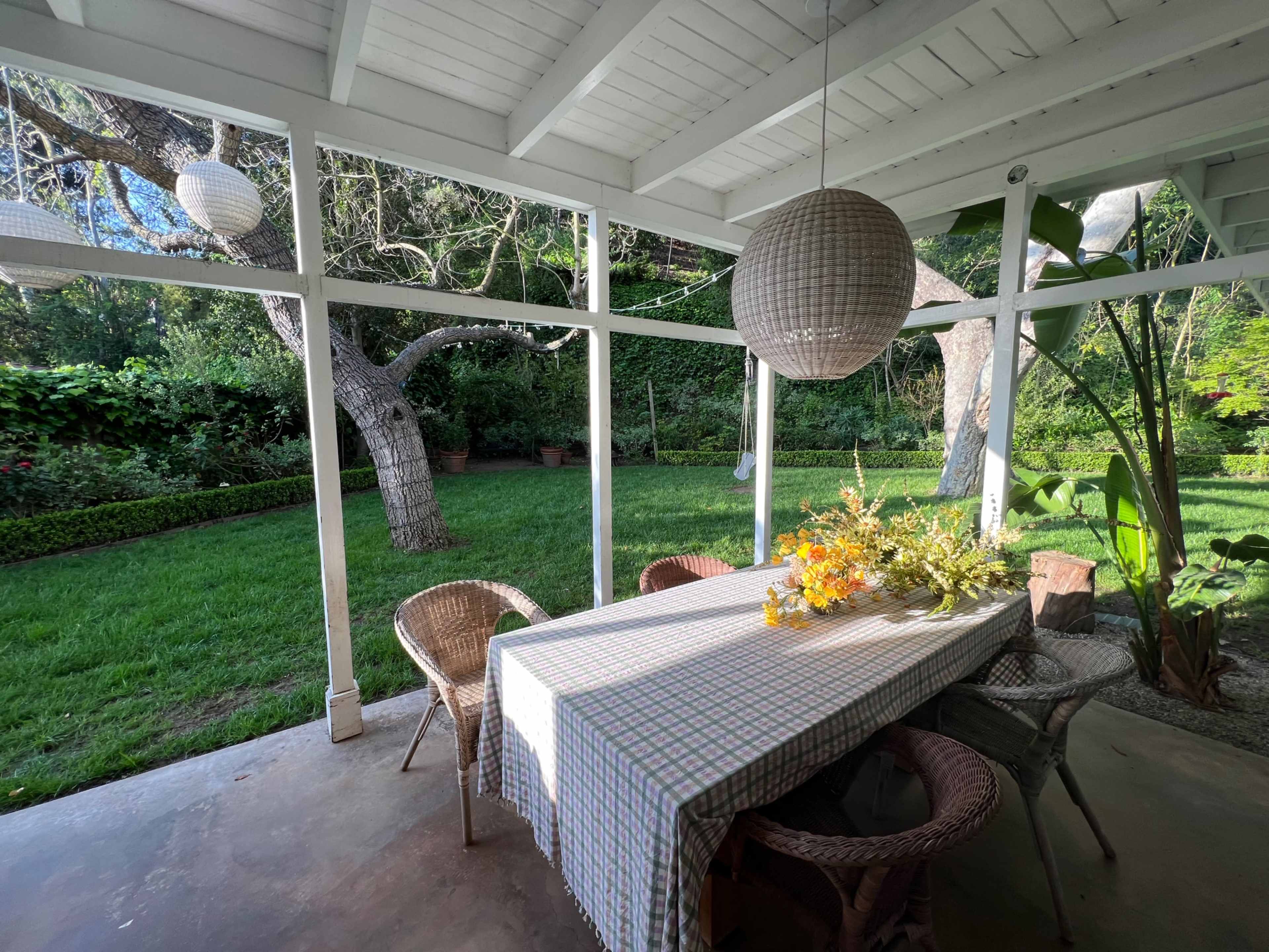 A shaded outdoor dining area features a table adorned with a floral centerpiece, surrounded by wicker chairs and framed by greenery.