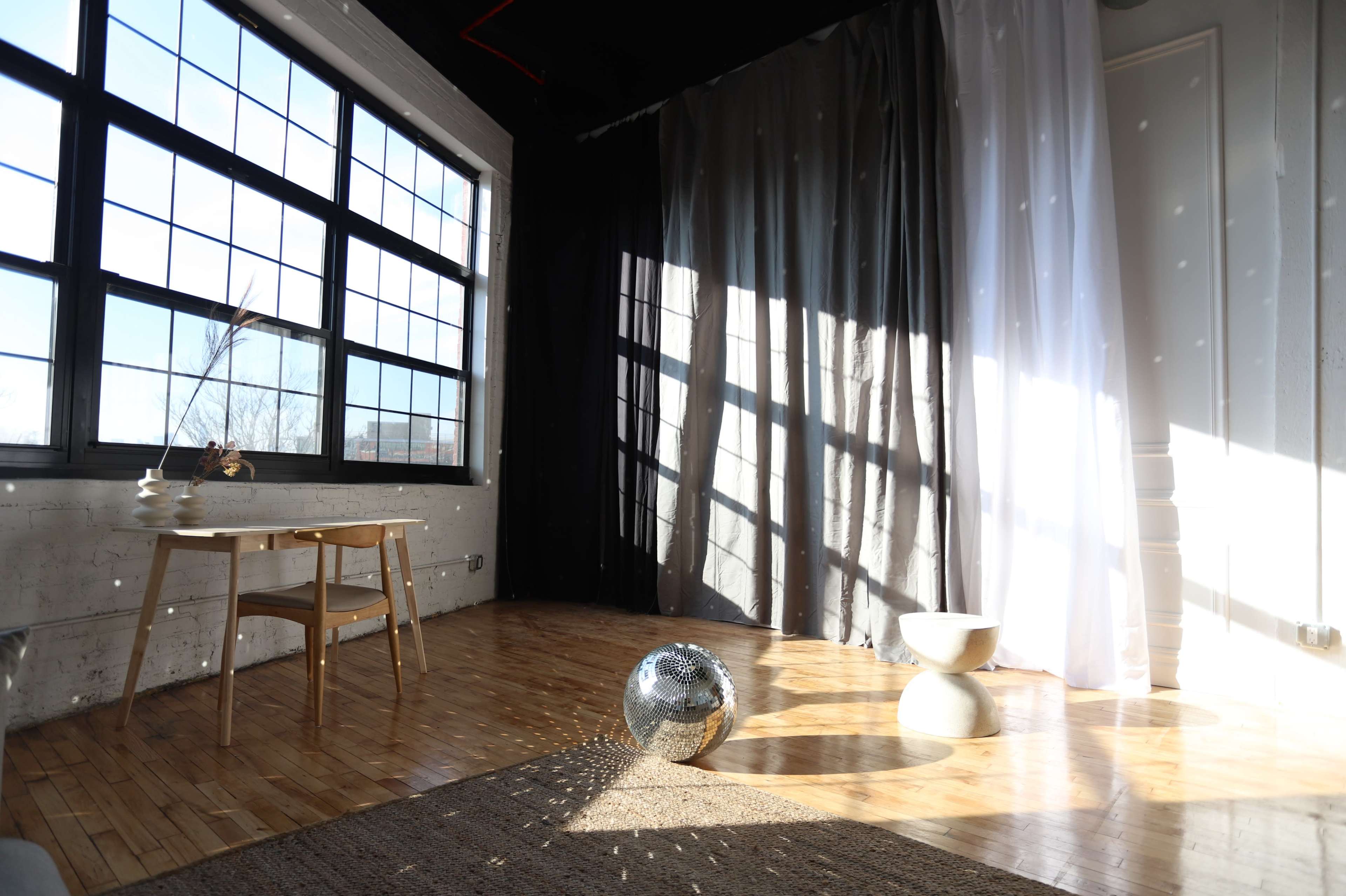 A well-lit room with large windows, a wooden floor, a round mirror ball, and minimalist furniture including a table and two decorative pieces.