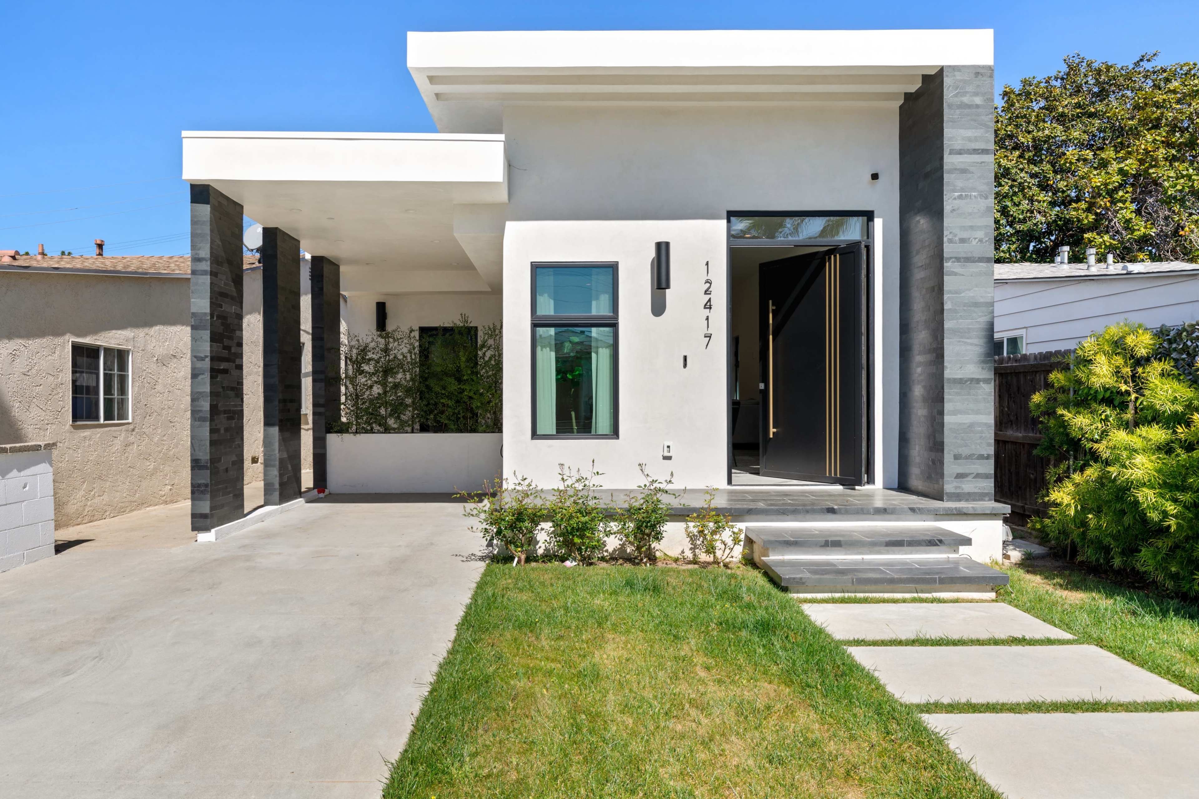 The image shows a modern, single-story house with a front porch, landscaped yard, and a pathway leading to the entrance.