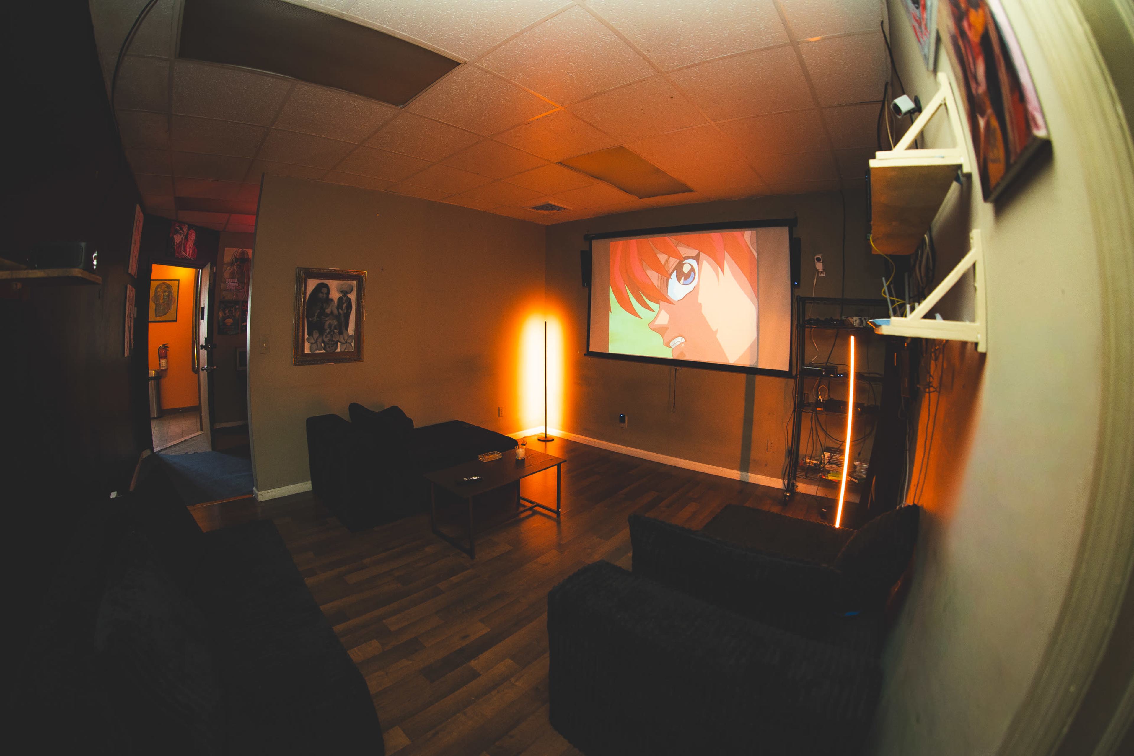 A cozy room features a projector screen displaying an animated character, with dark couches and ambient lighting.