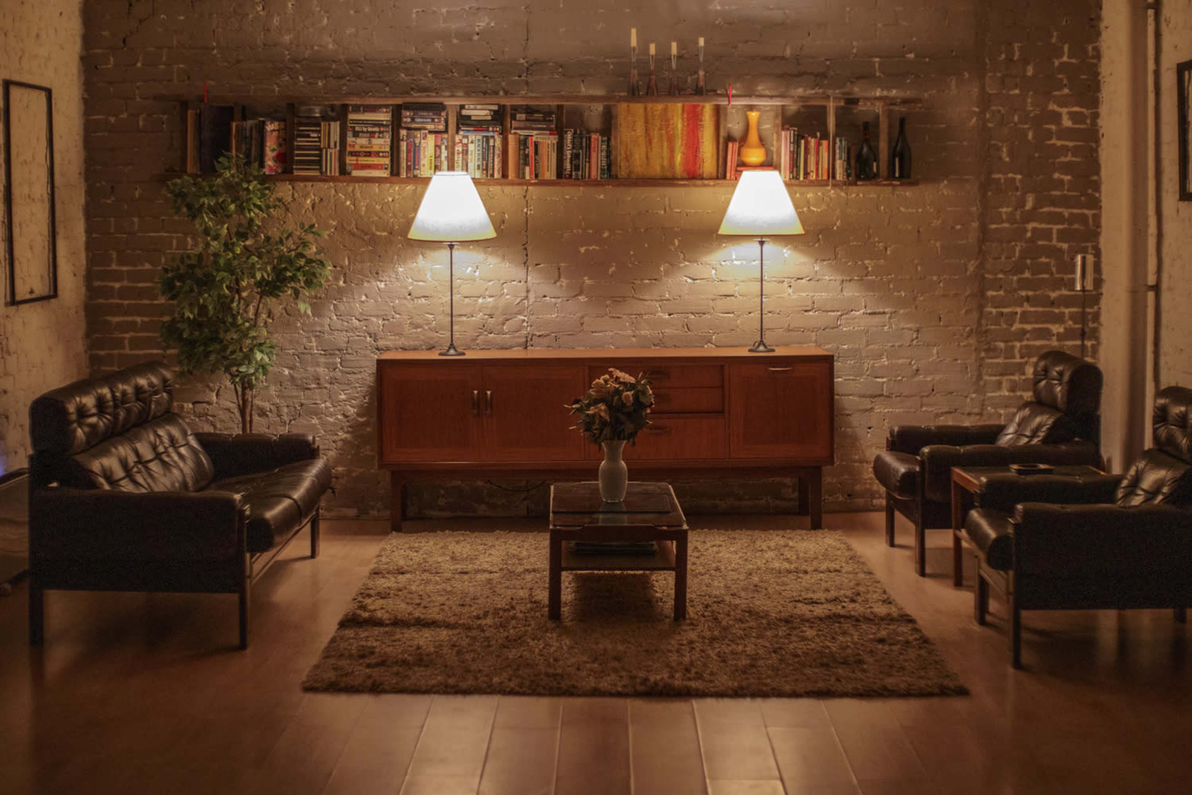 A cozy living room with two leather sofas, a wooden cabinet, a coffee table, and two lamps providing soft lighting, surrounded by a bookshelf and a potted plant.