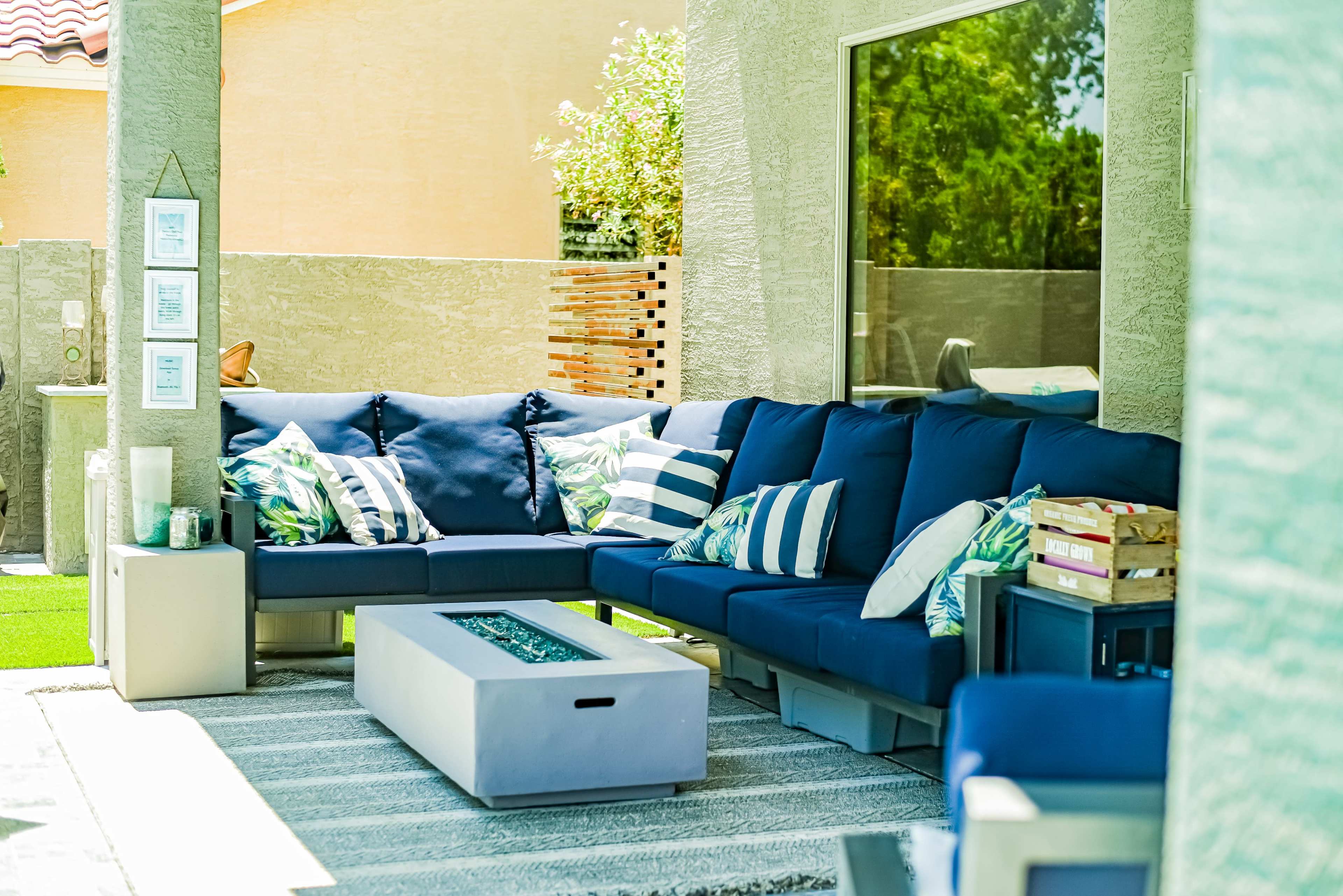 A modern outdoor seating area features a navy sectional sofa with decorative cushions, a rectangular fire table, and a textured rug.