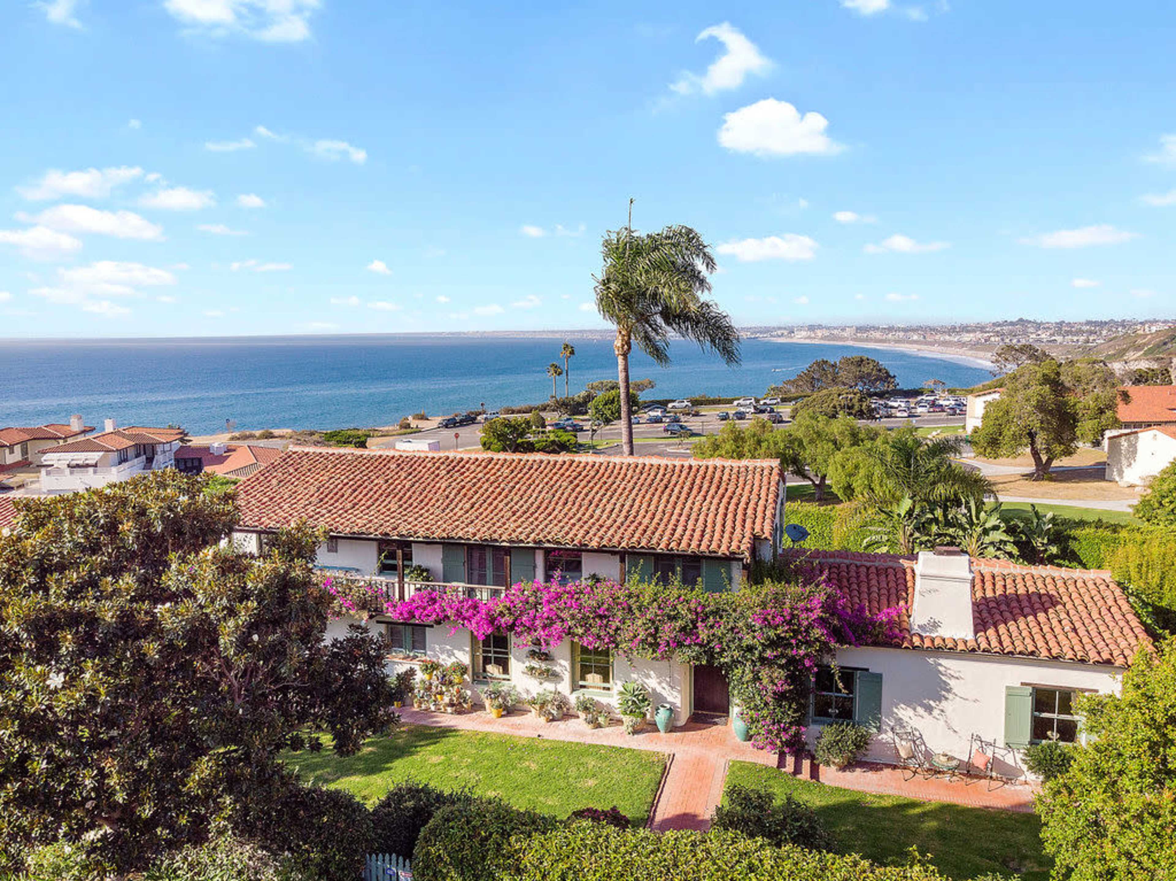 A two-story house with a red tiled roof is surrounded by lush greenery and features purple flowers, overlooking a coastal view.