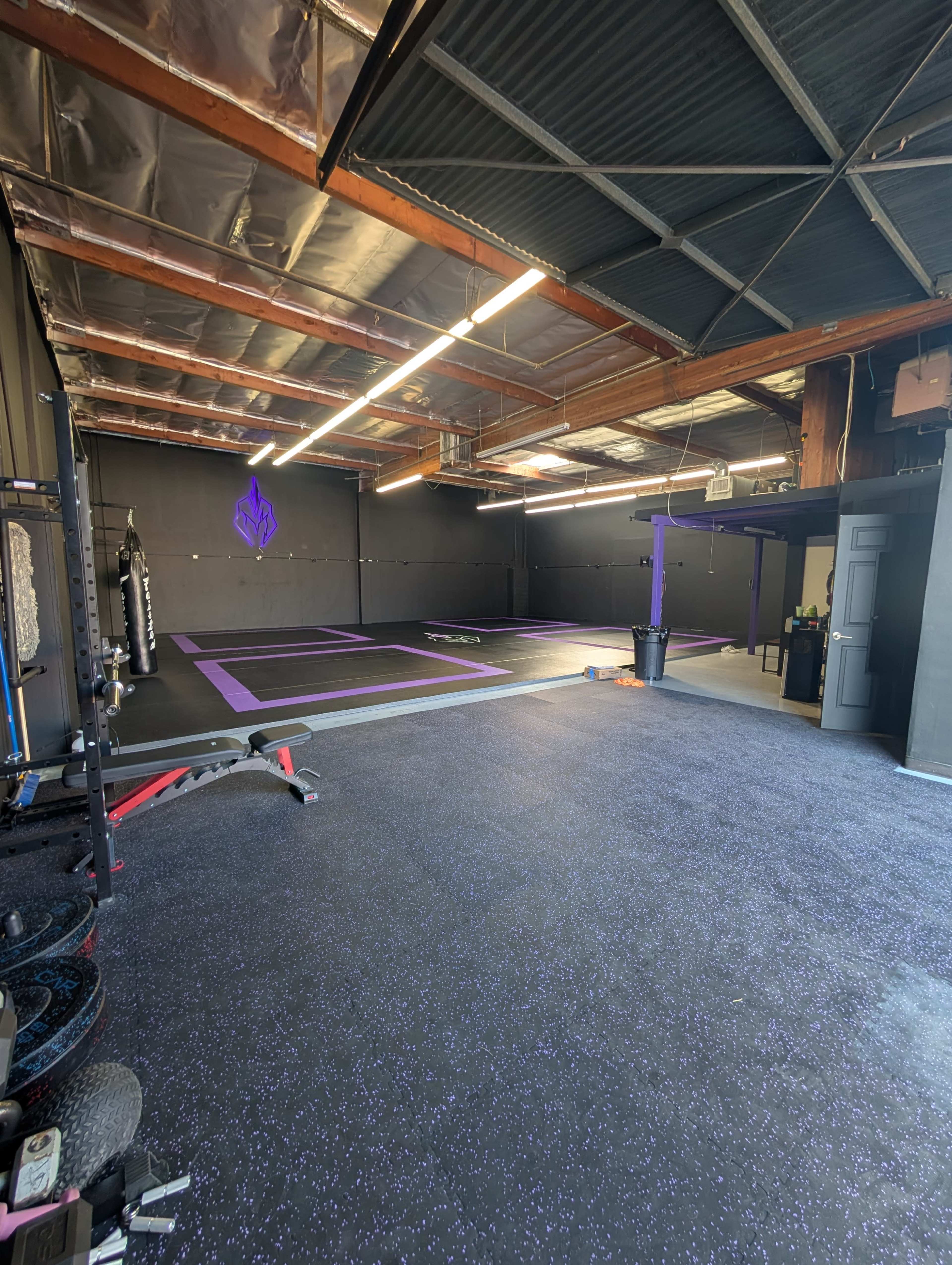 The image shows a spacious gym with black walls, purple flooring, and various exercise equipment along one side.