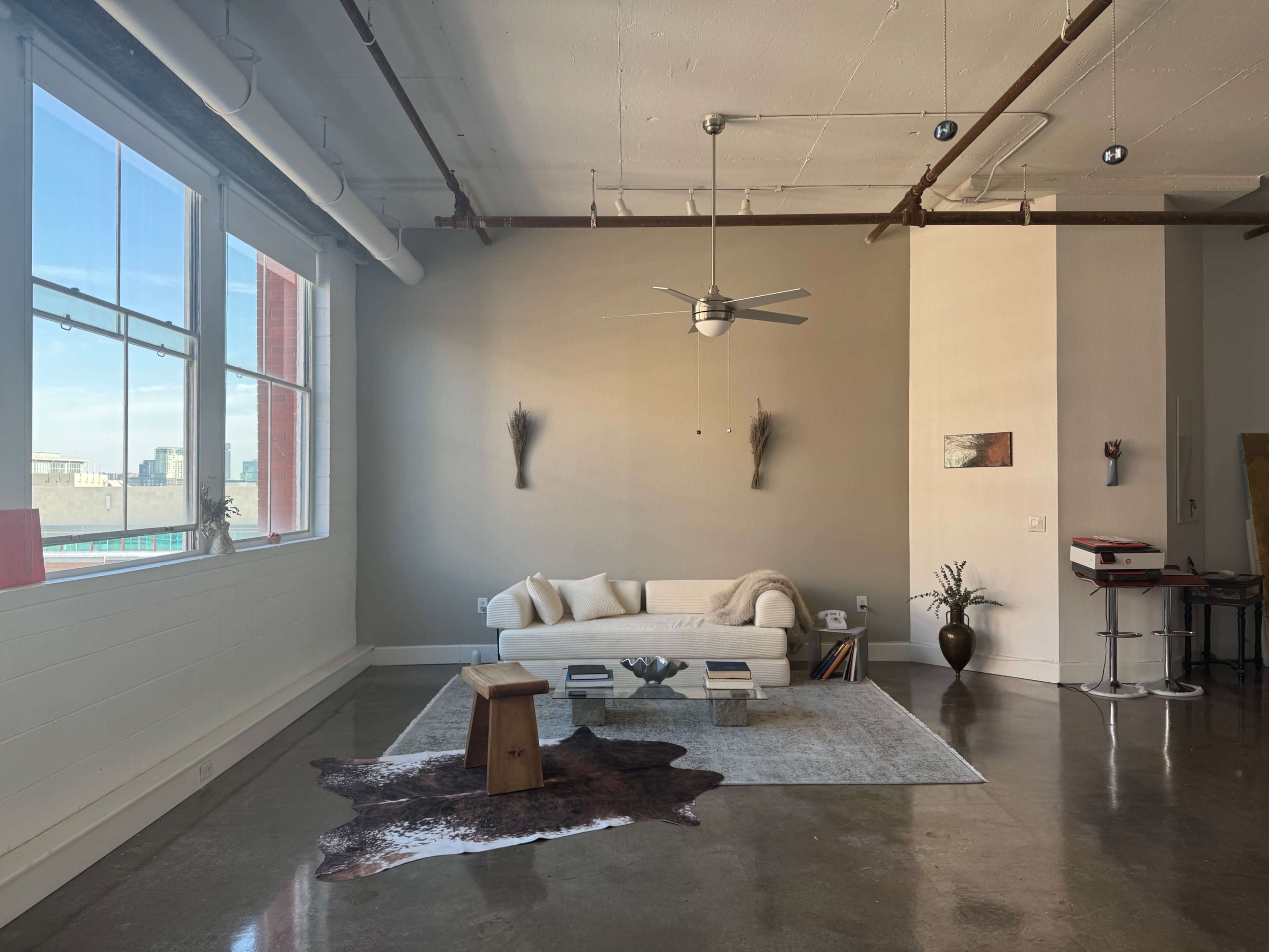 The image shows a spacious, modern living area with large windows, a light-colored sofa, a coffee table, and a decorative rug on polished concrete flooring.