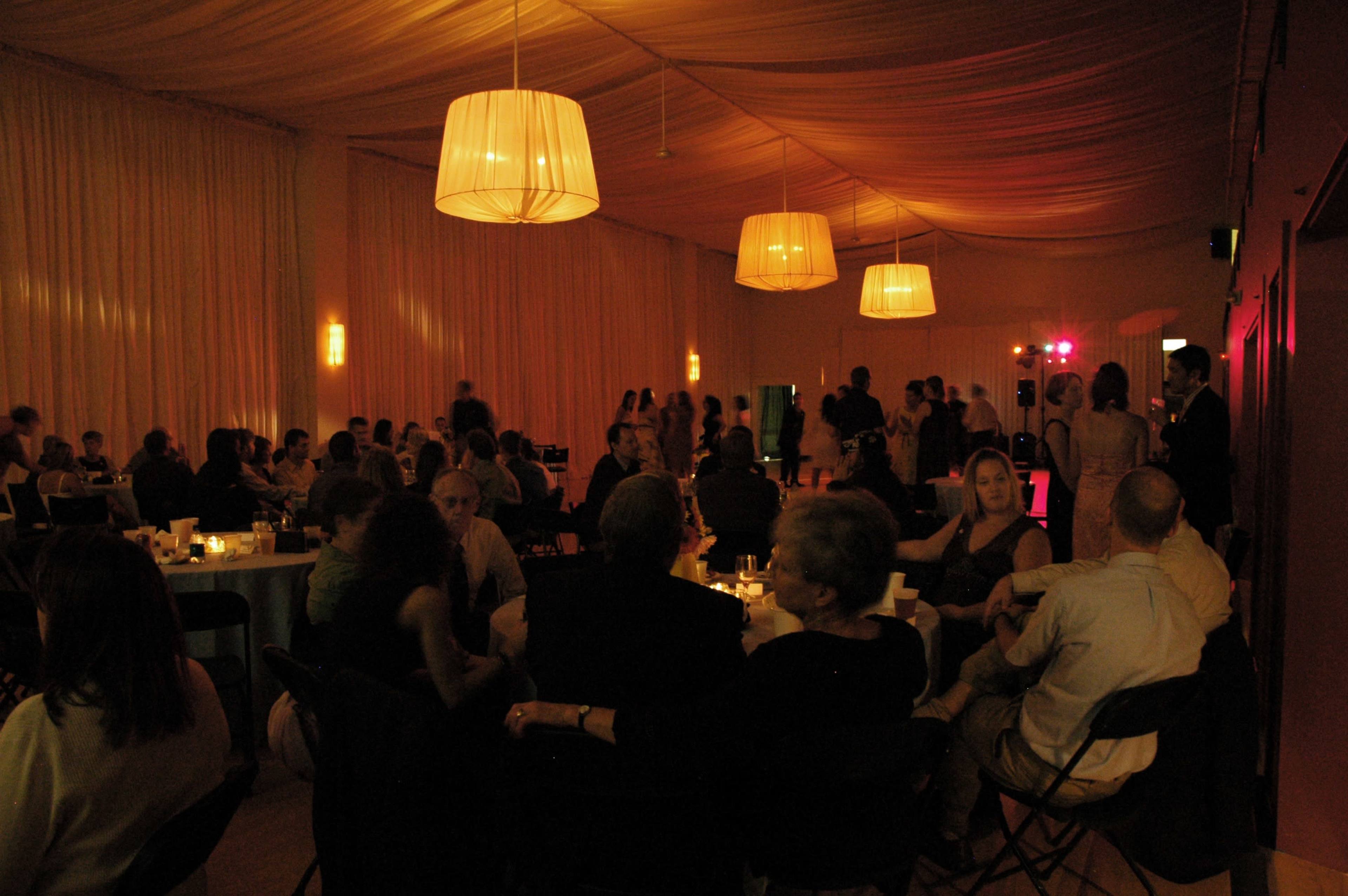 A large indoor event space is filled with tables of guests, while a dance floor is illuminated by colorful lights and a DJ setup in the background.