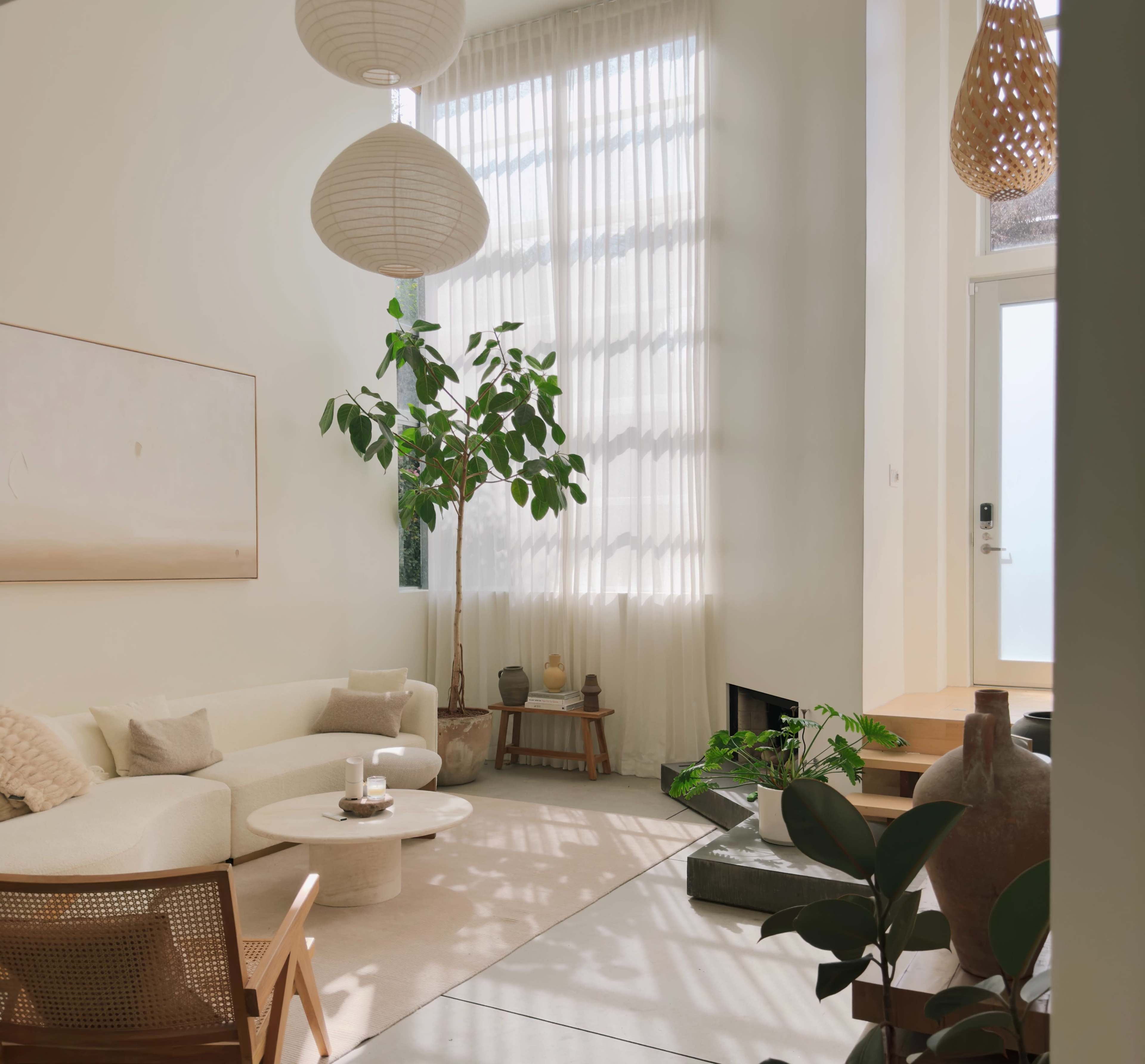 A bright living room features a white sectional sofa, a round coffee table, large windows with sheer curtains, and various indoor plants.