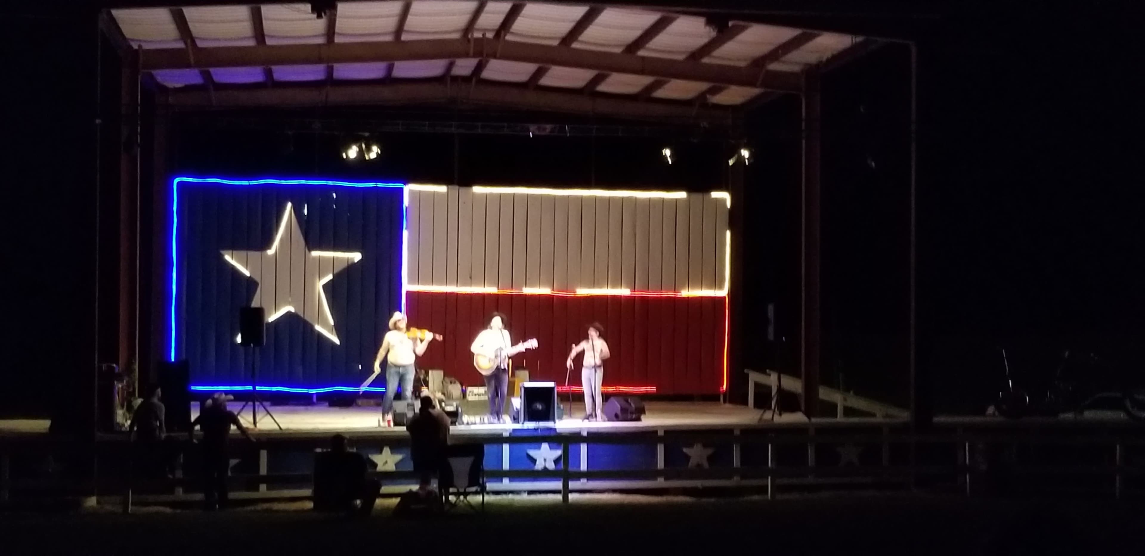 A band performs on stage illuminated by a backdrop featuring a large Texas flag.