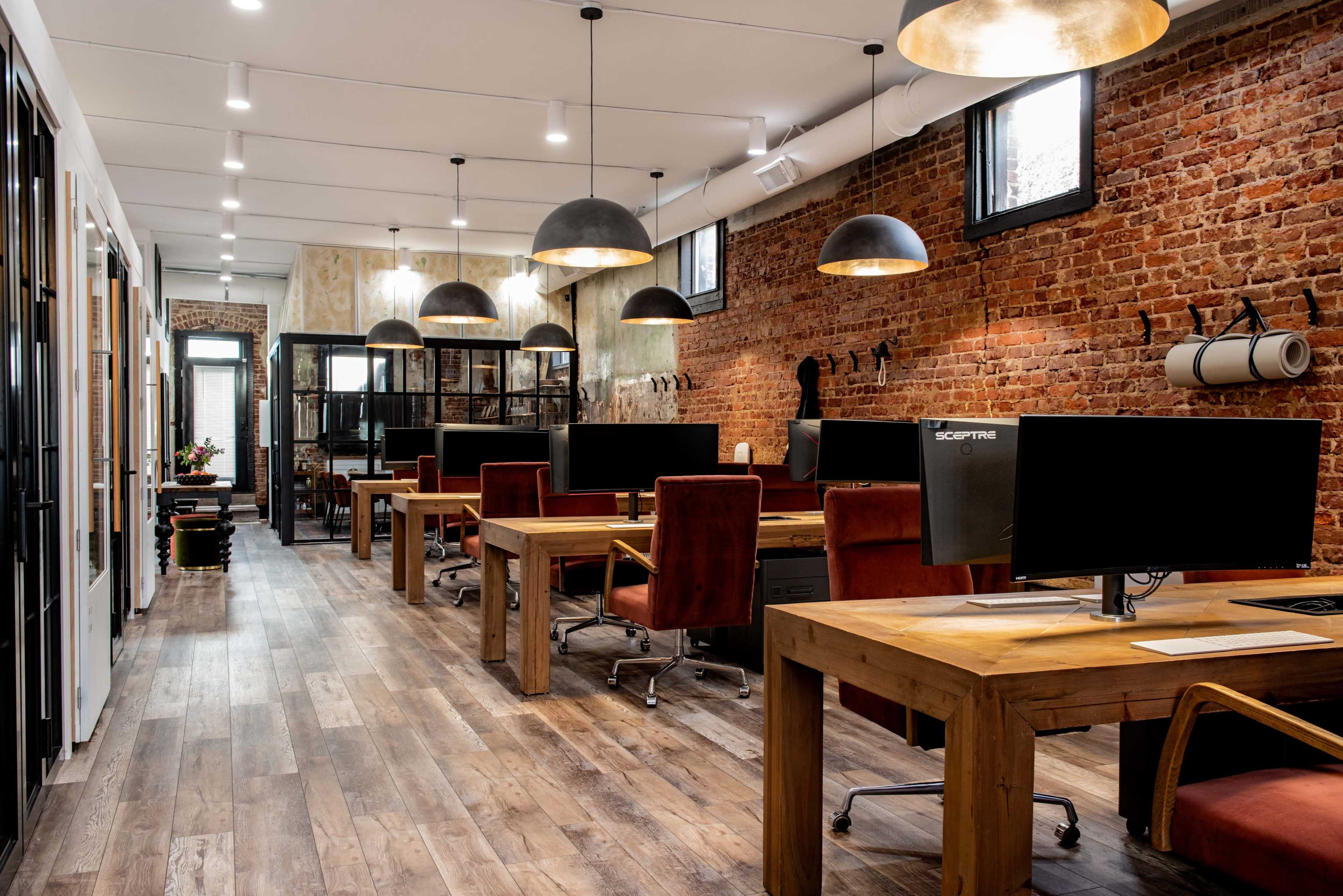 A modern office space with wooden desks, large computer monitors, and an exposed brick wall, illuminated by pendant lights.