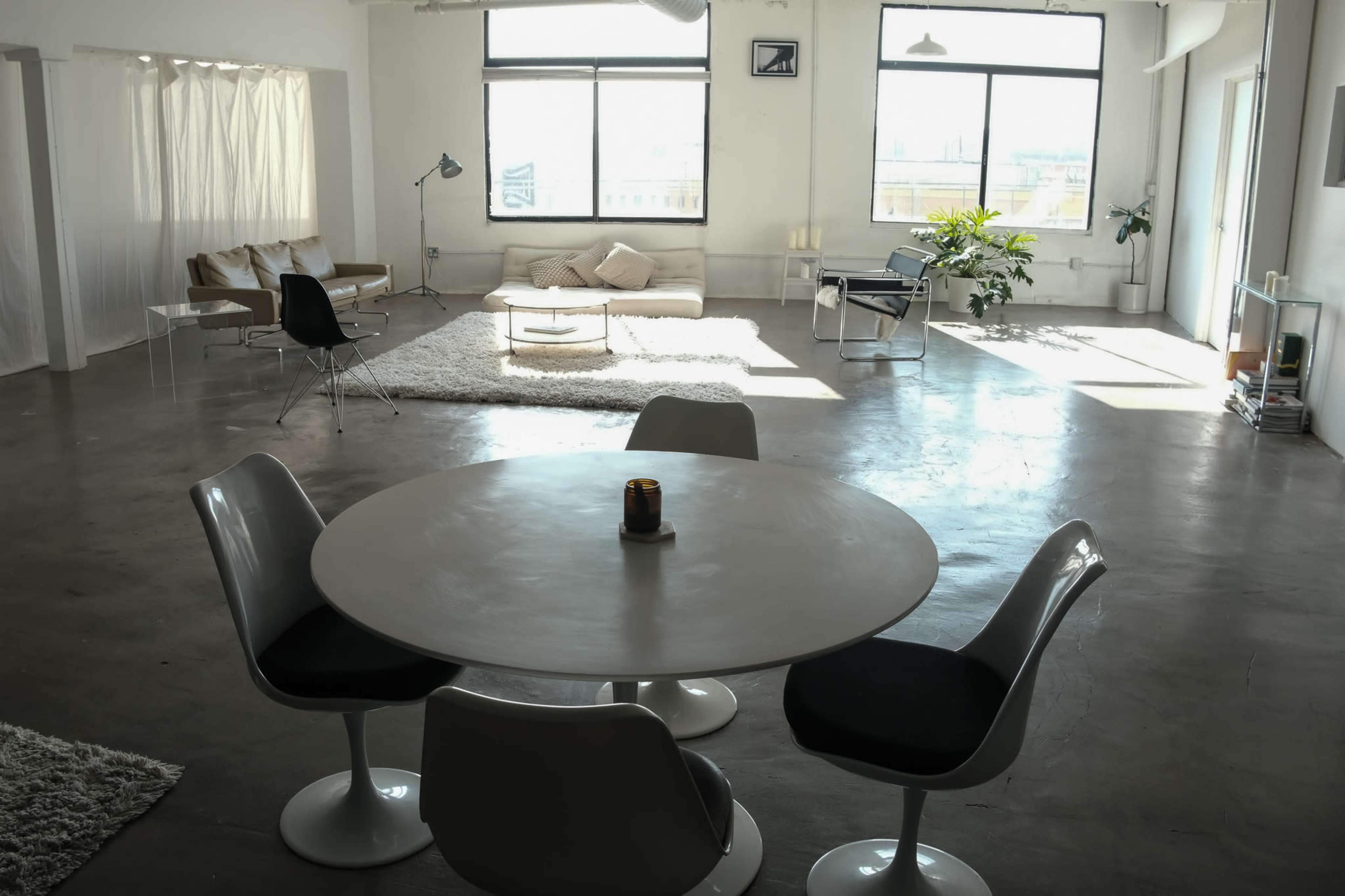The image shows a spacious, modern interior with a round dining table and chairs in the foreground, alongside a living area featuring a couch, plants, and large windows that allow natural light to fill the space.