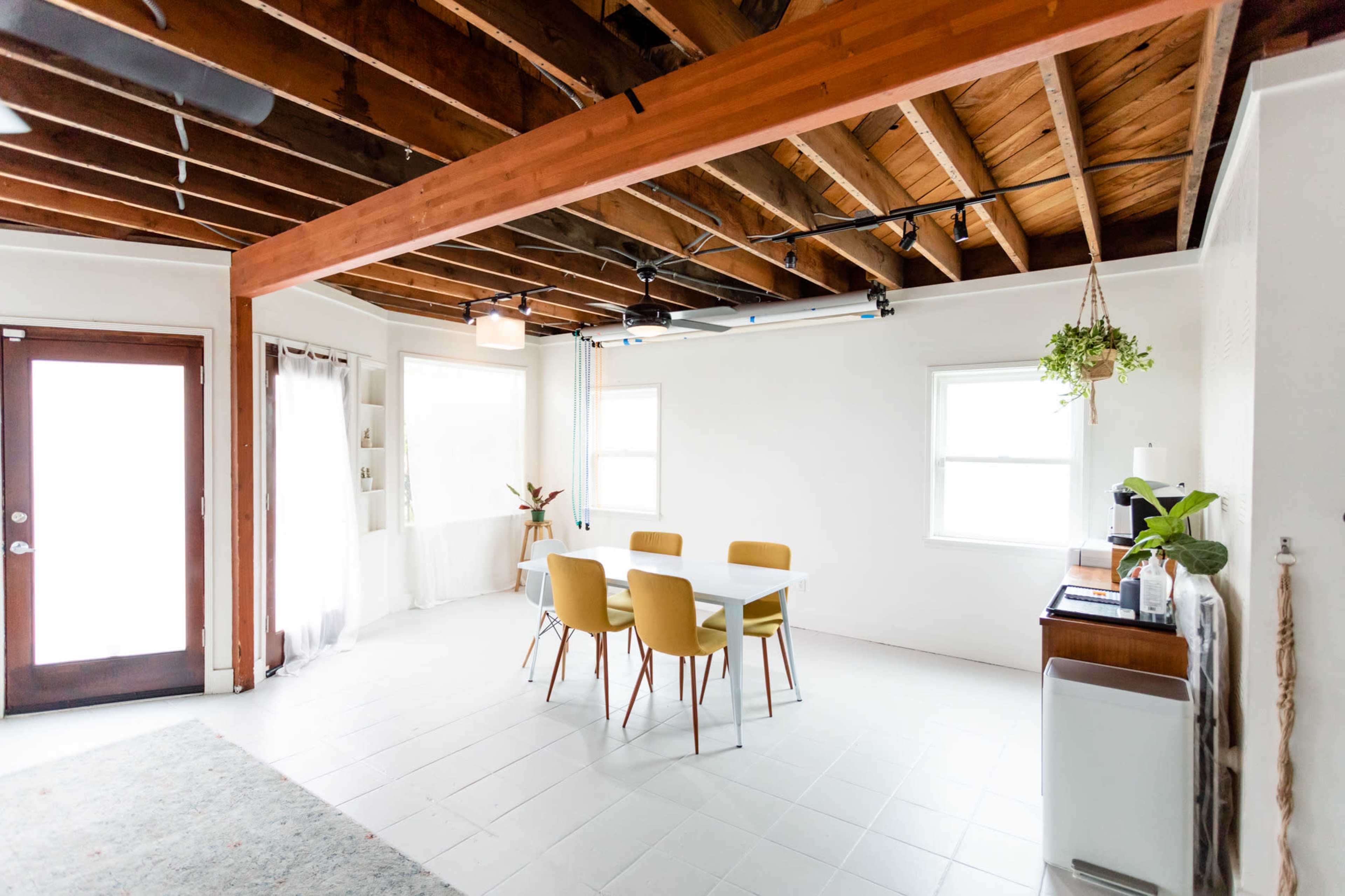 A bright, minimalist dining area features a white table surrounded by yellow chairs beneath exposed wooden beams.