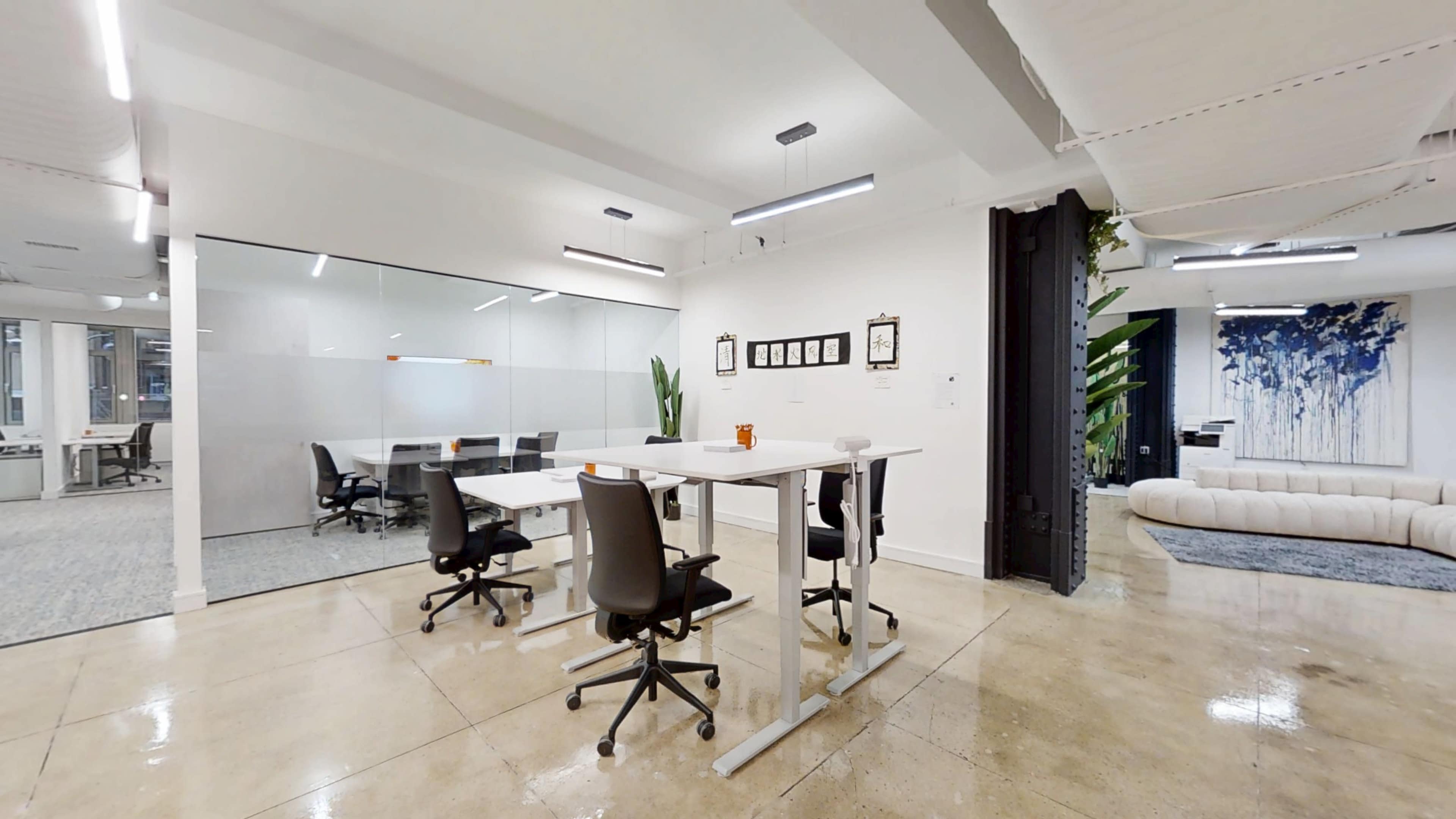 The image shows a bright, modern office space featuring a communal table with chairs, a glass-walled meeting room, and decorative plants.