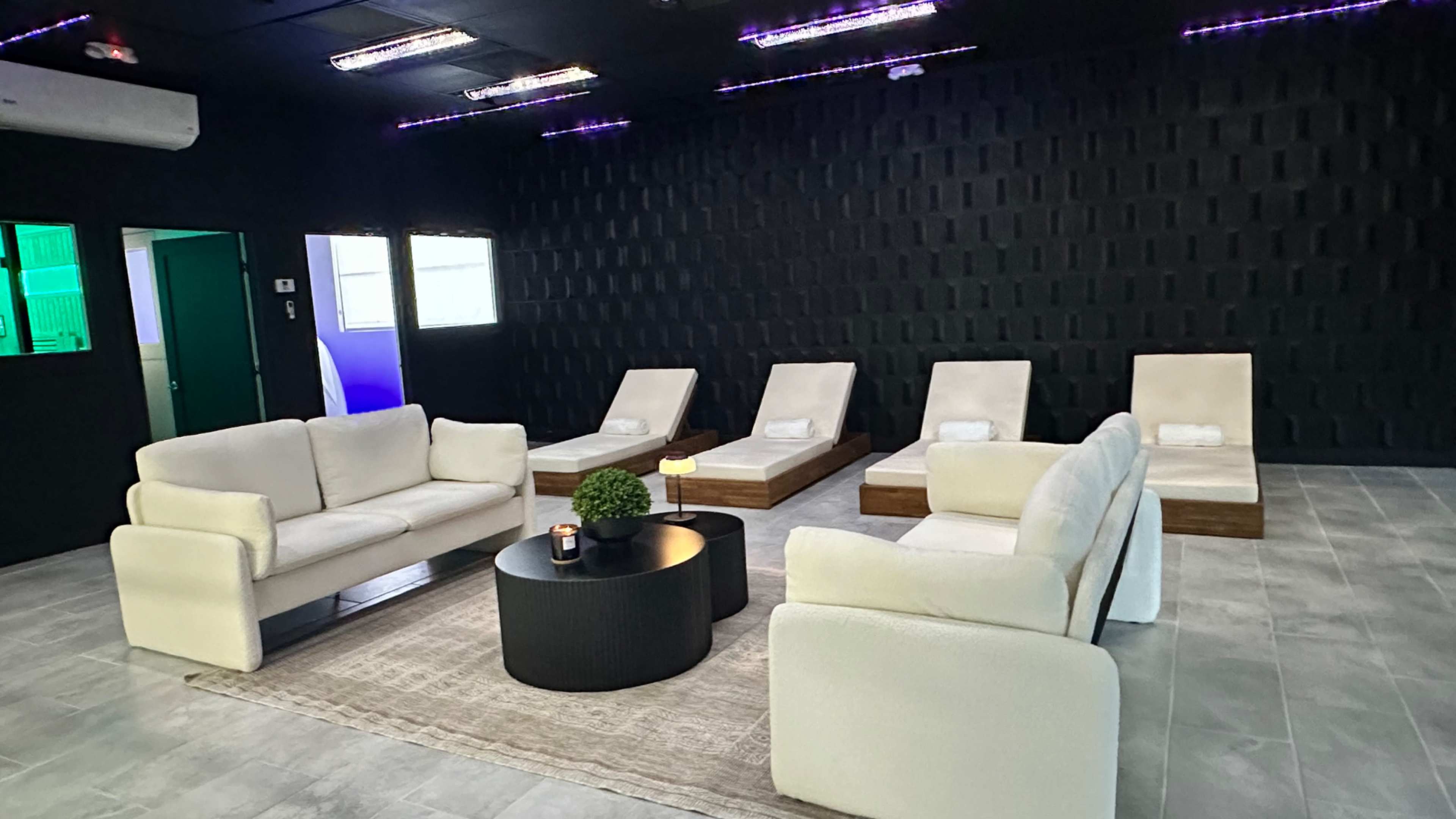 A modern relaxation room with two white sofas, a round black coffee table, and several lounge chairs arranged around a cozy area rug.