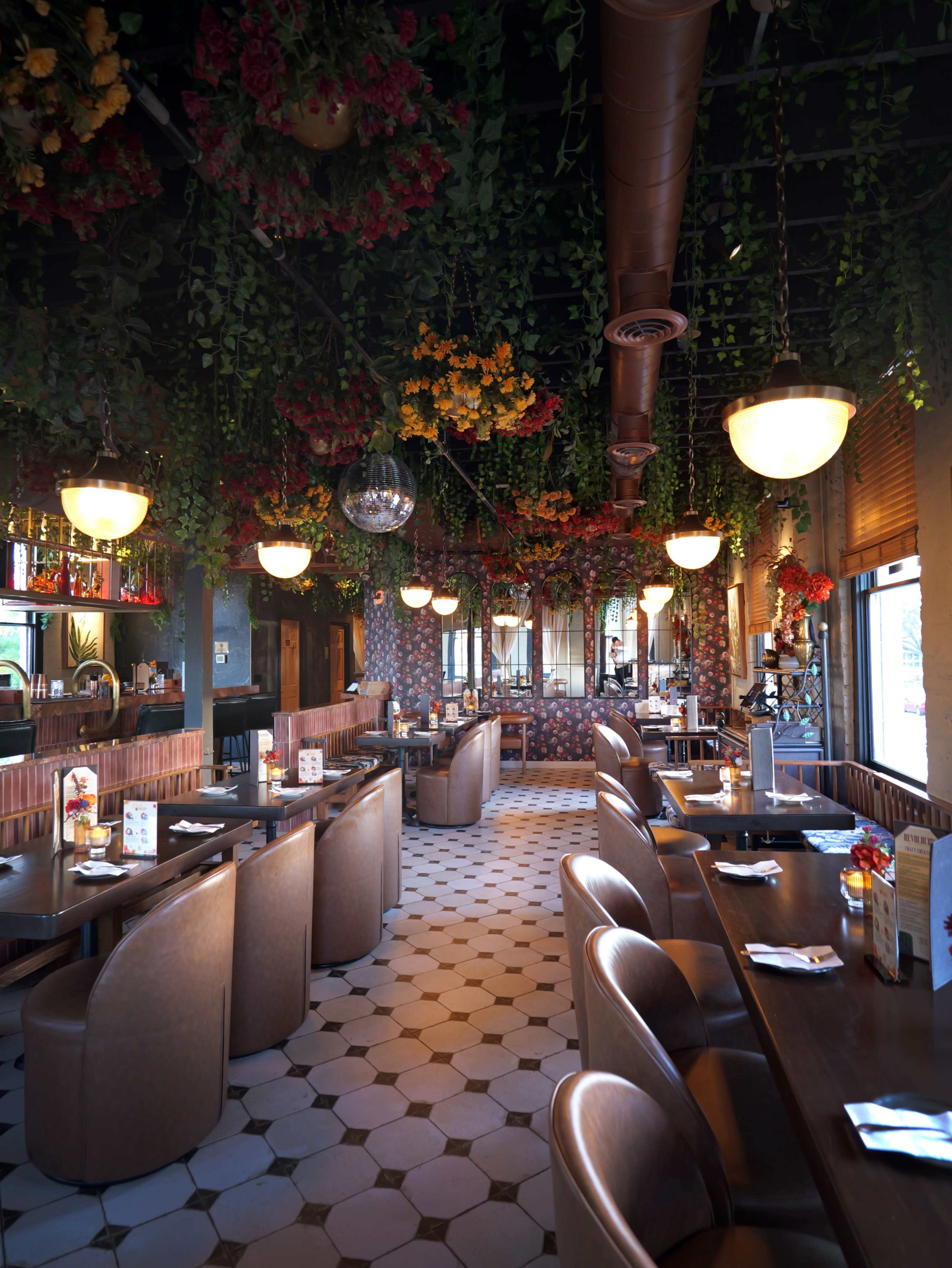 The image shows a restaurant interior with floral decorations hanging from the ceiling, round pendant lights, and tables arranged along the walls.