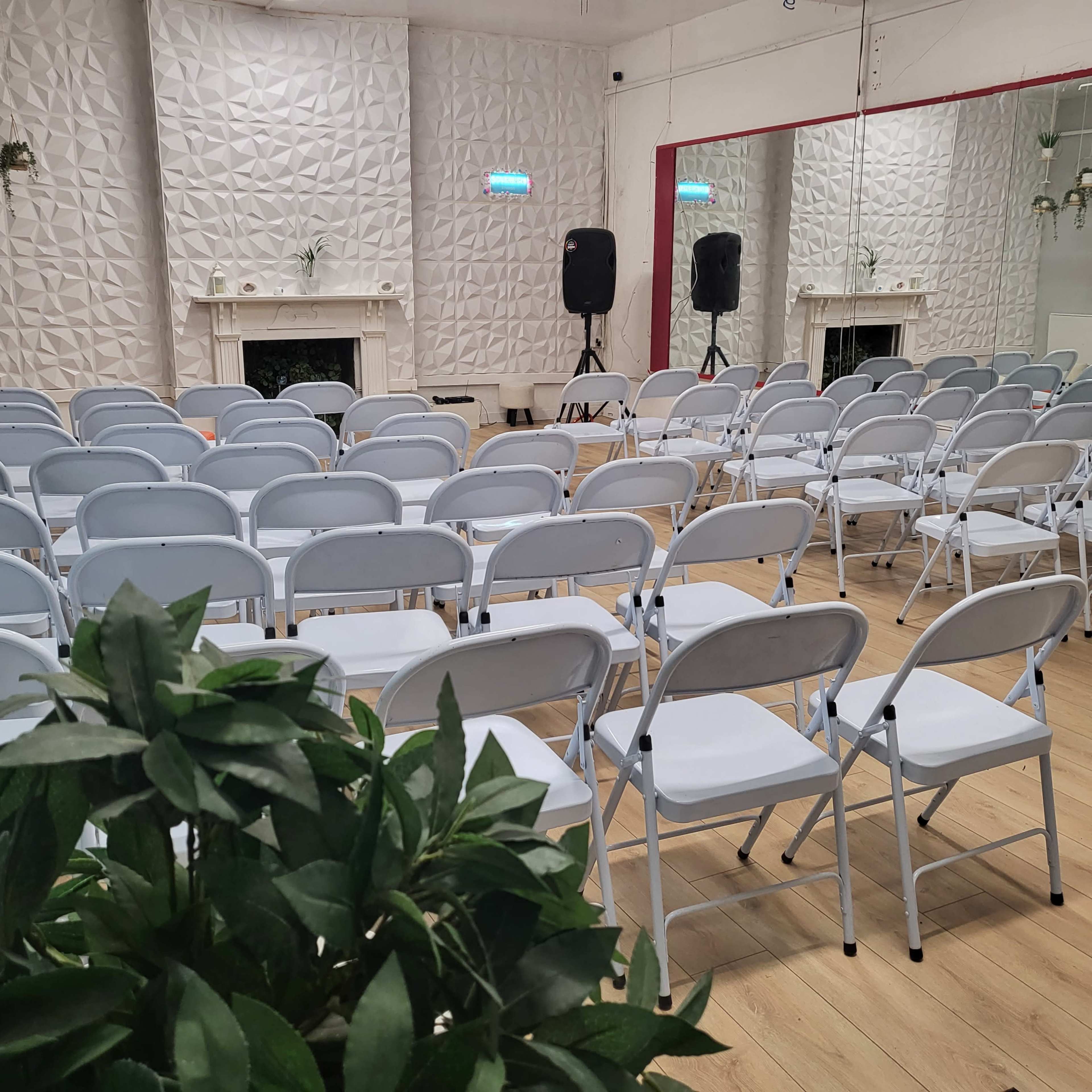 The image shows a seminar room filled with rows of white folding chairs facing a mirrored wall, with a decorative fireplace and two speakers in the background.