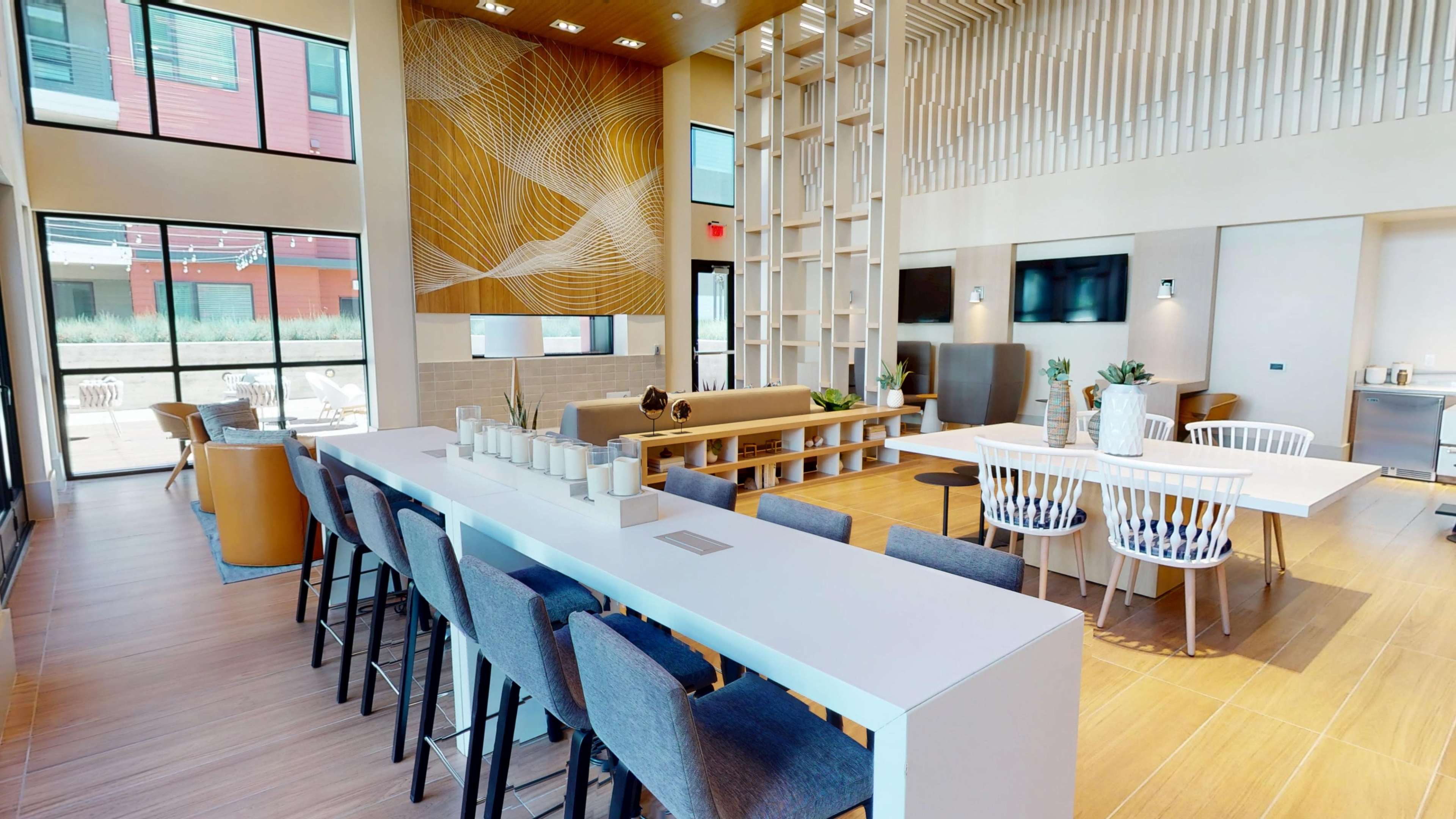 The image depicts a modern communal lounge area featuring a long white table surrounded by chairs, a cozy seating arrangement, and large windows that allow natural light to fill the space.