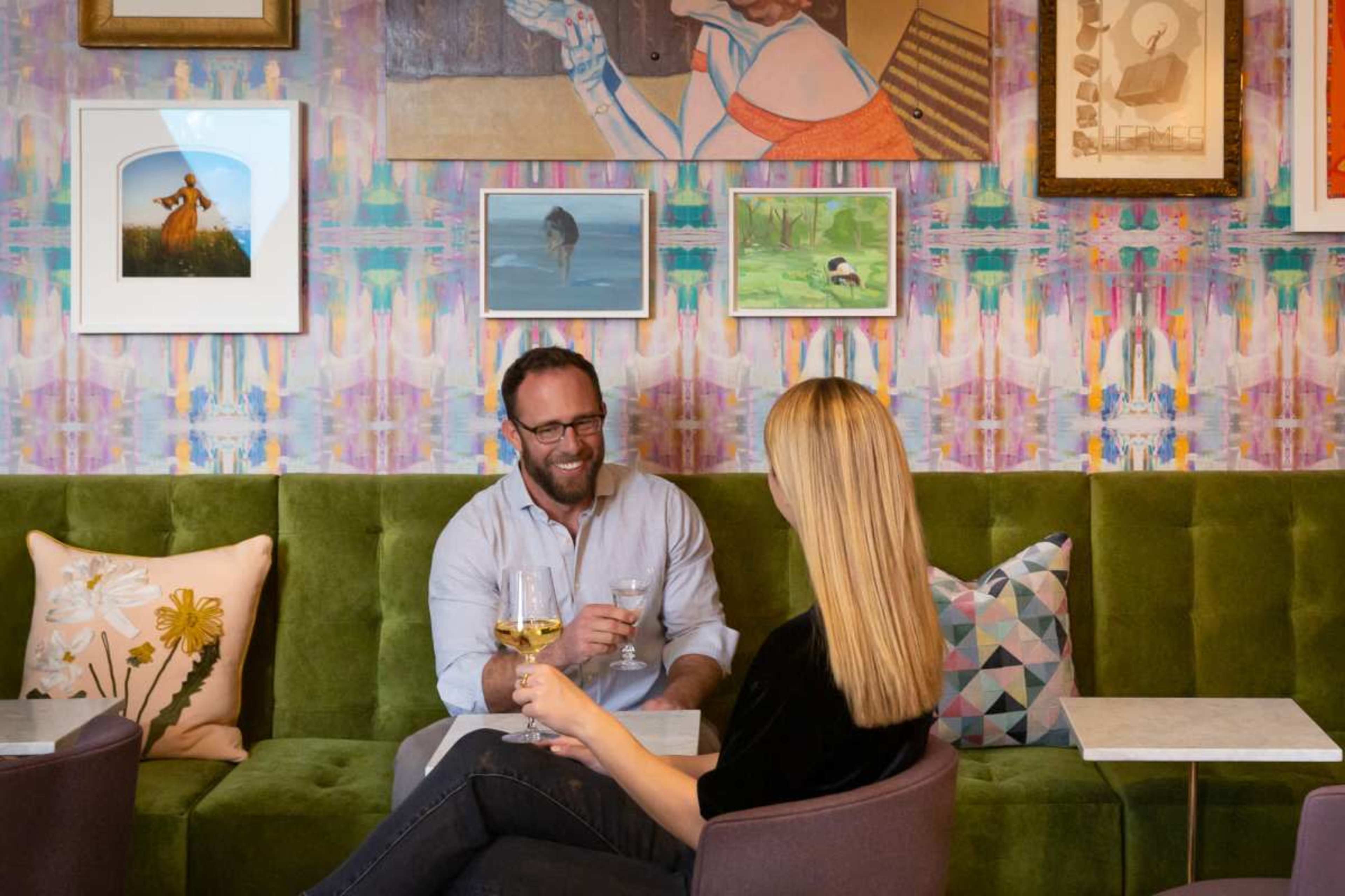 A man and a woman are seated on a green couch in a colorful restaurant, toasting with glasses of wine in front of a wall adorned with various art pieces.