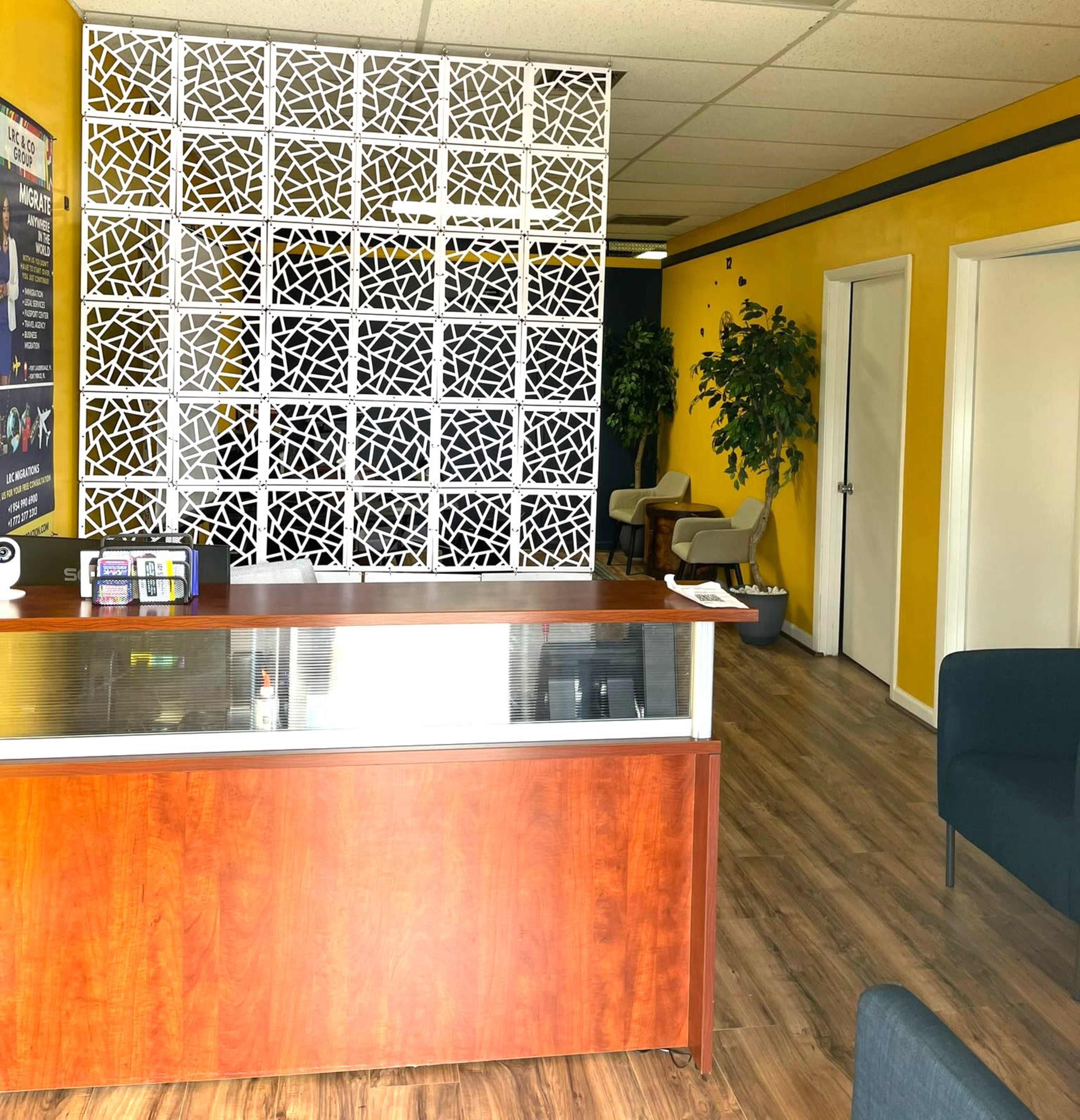 A reception area with a wooden front desk, a decorative partition, and a yellow wall, leading to a waiting area with seating.