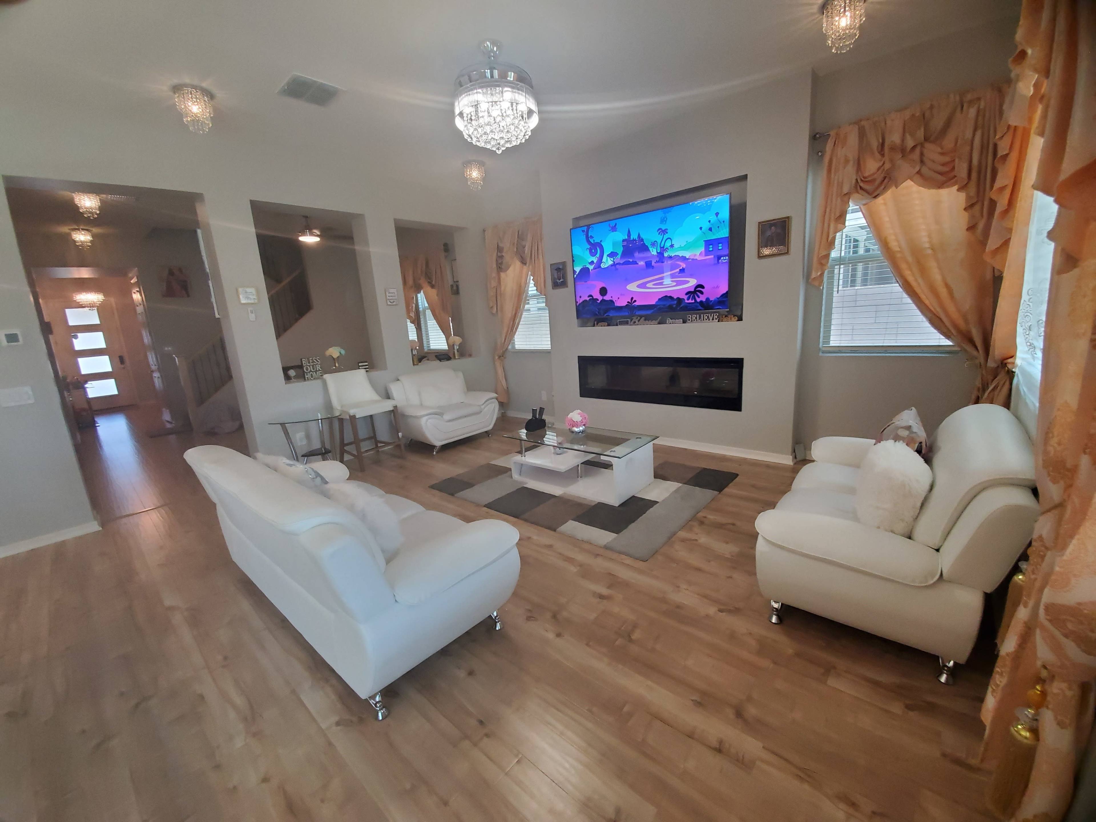 The living room features two white sofas, a glass coffee table, and a large wall-mounted TV, with decorative curtains and a modern fireplace.