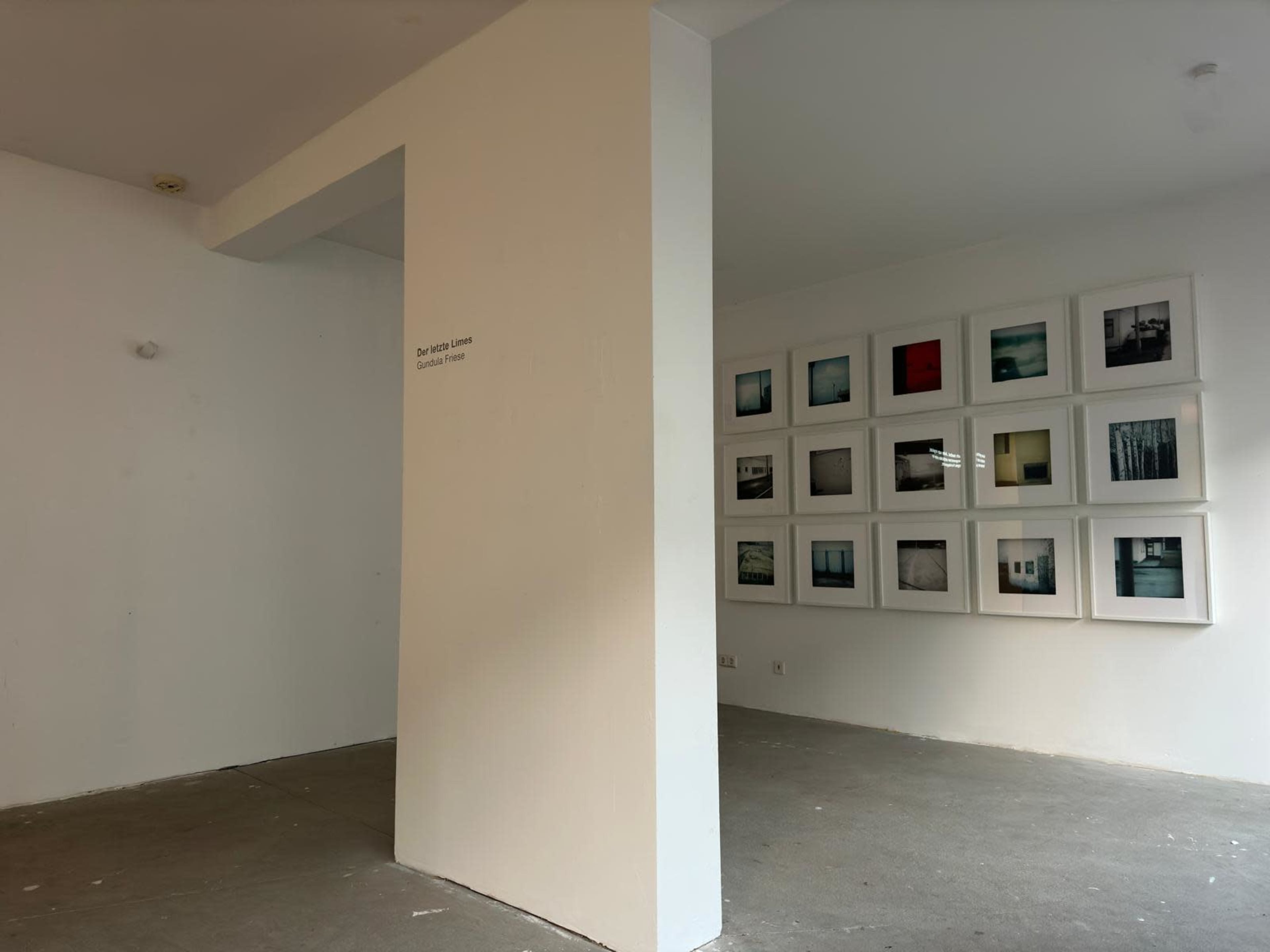 The image shows a partially painted white room with a blank wall featuring a series of framed photographs arranged in a grid on the opposite wall.