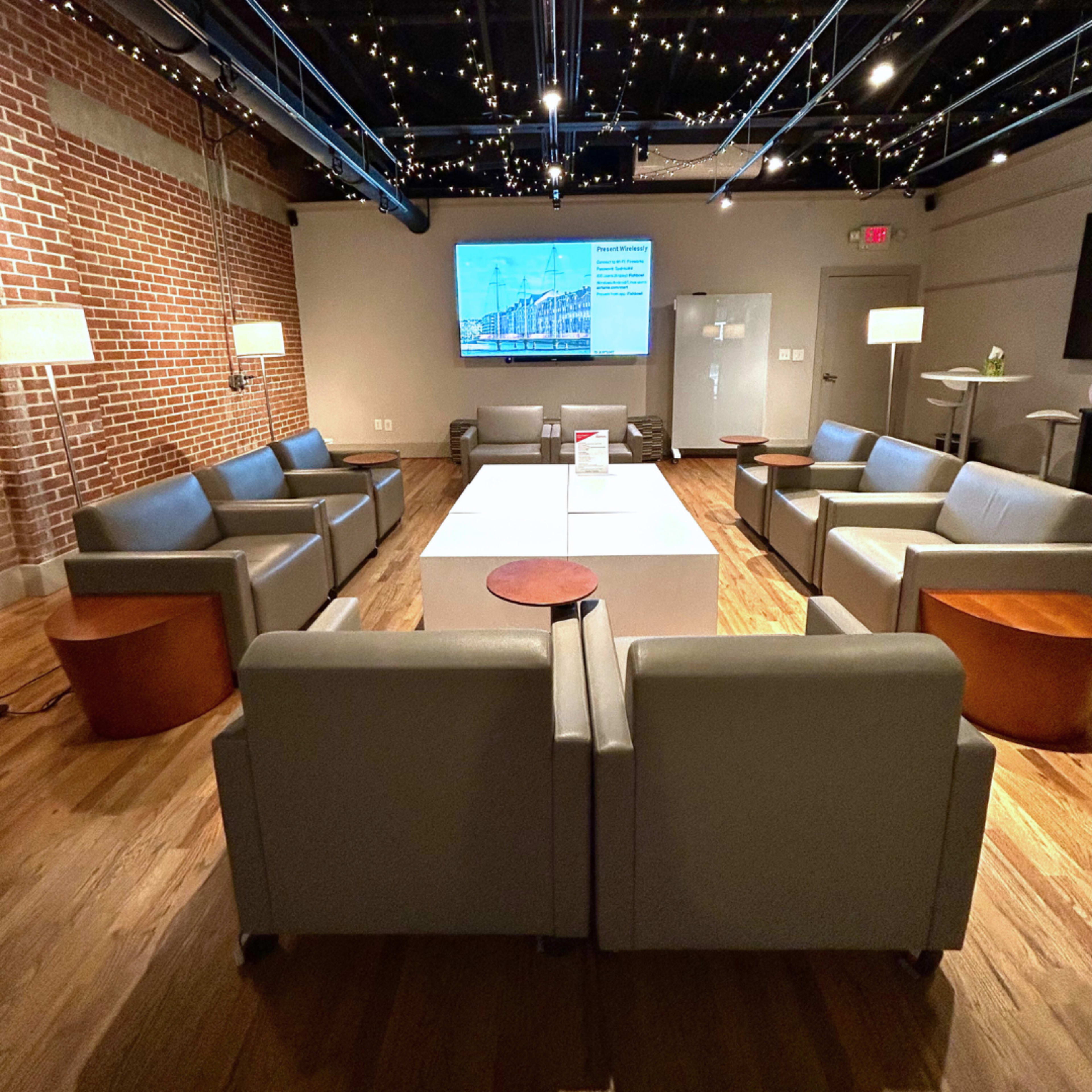 A modern lounge area features several gray sofas arranged around a central white table, with wooden side tables and a large screen displaying information on the wall.