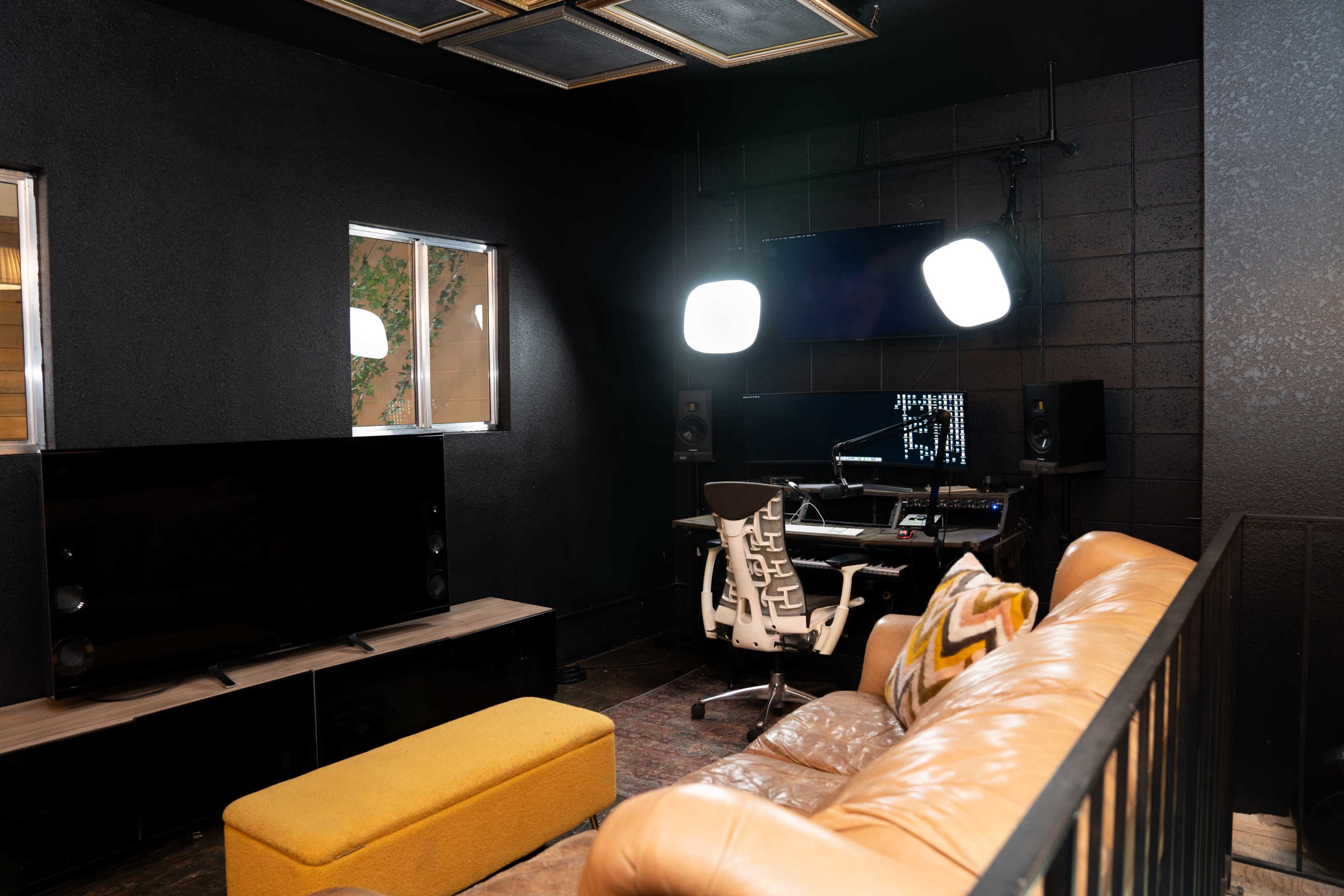 A modern recording studio with a computer workstation, black walls, a brown leather couch, and a yellow ottoman.